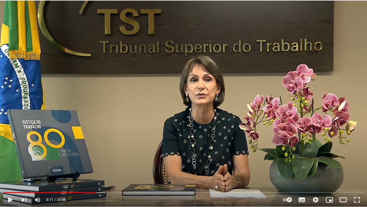 Ministra Maria Cristina Peduzzi no lançamento do livro de 80 Anos da Justiça do Trabalho, ela é branca, está de blusa preta com bolinhas douradas, o cabelo dela é curso não chegando ao ombro, as mãos estão juntas em cima da mesa, na mesa há cópias do livro e um jarro com flores cor-de-rosa, atrás dela há uma bandeira do brasil e uma placa do tribunal superior do trabalho