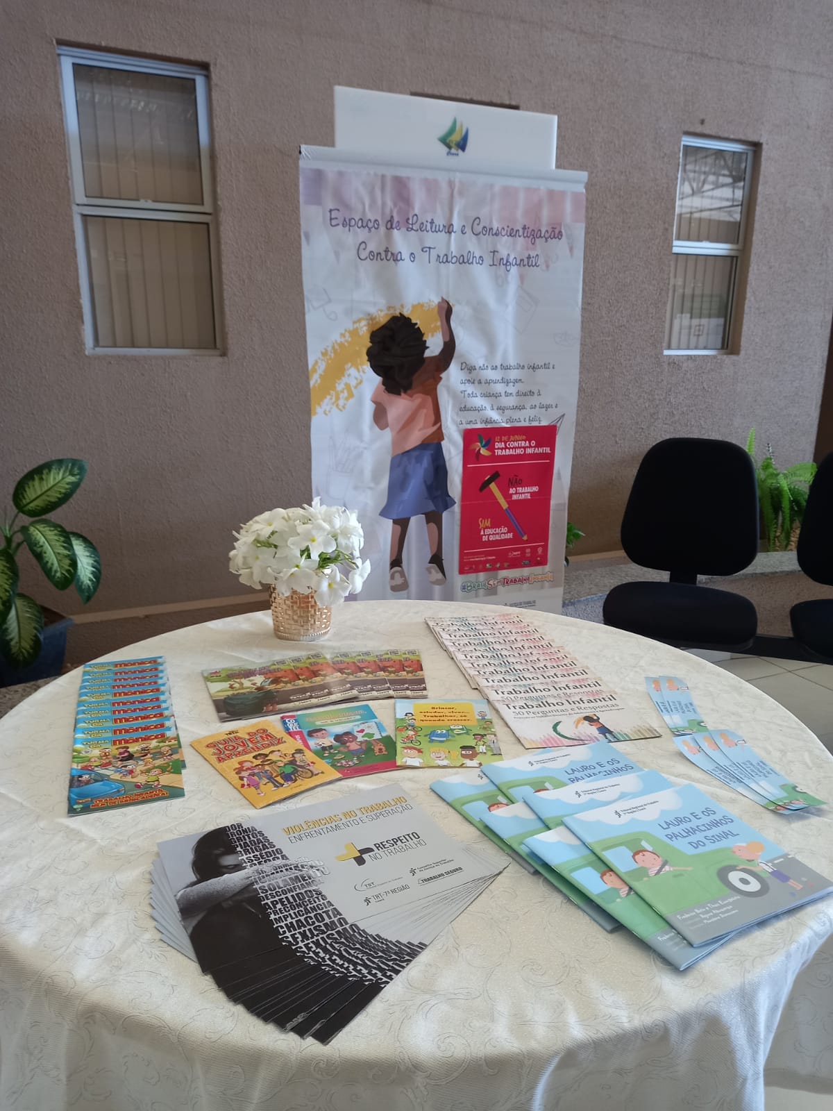 Imagem de espaço no Fórum do Cariri com material do Programa de Combate ao Trabalho Infantil em cima de uma mesa (livros, cartilhas) e um banner ao fundo