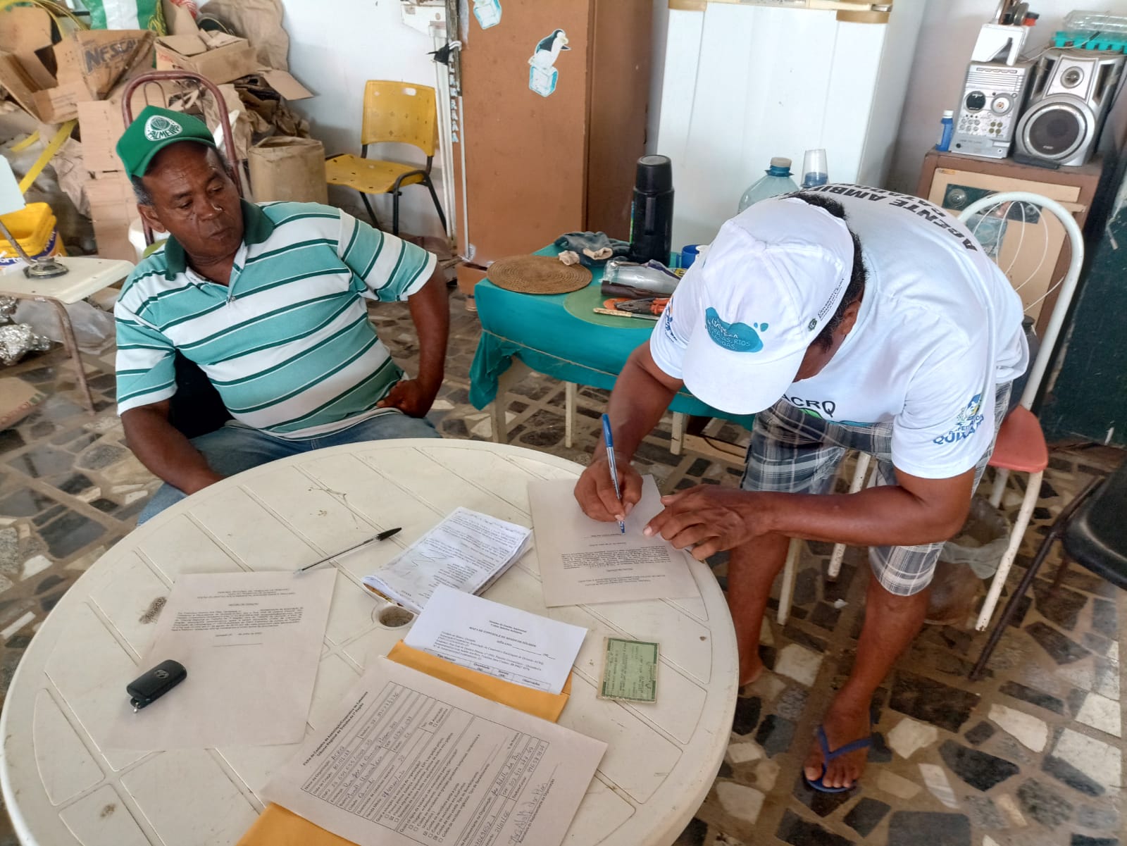 Foto de dois integrantes de associação de catadores assinando documentos. ao redor tem materiais descartáveis