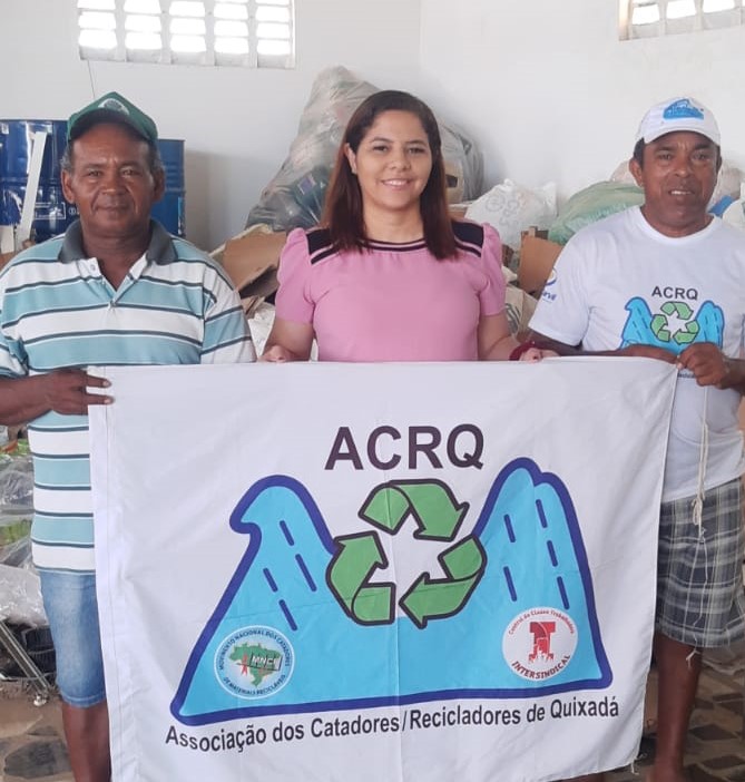 Foto da diretora de secretaria de Quixadá, Geyza Moura e representantes da Associação de Catadores de Quixadá segurando uma bandeira com a logo e nome da Associação