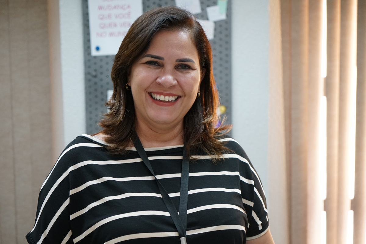 Foto da assistente social Rosiane Barros. Ela está com blusa listrada branca e preta, usa crachá e está sorrindo.