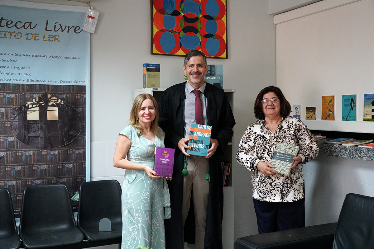 Foto Bibliotecária Rejane Albuquerque, juiz titular Jaime Luís e a presidente do TRT7, Regina Gláucia. Todos estão segurando um livro e estão em frente à biblioteca livre