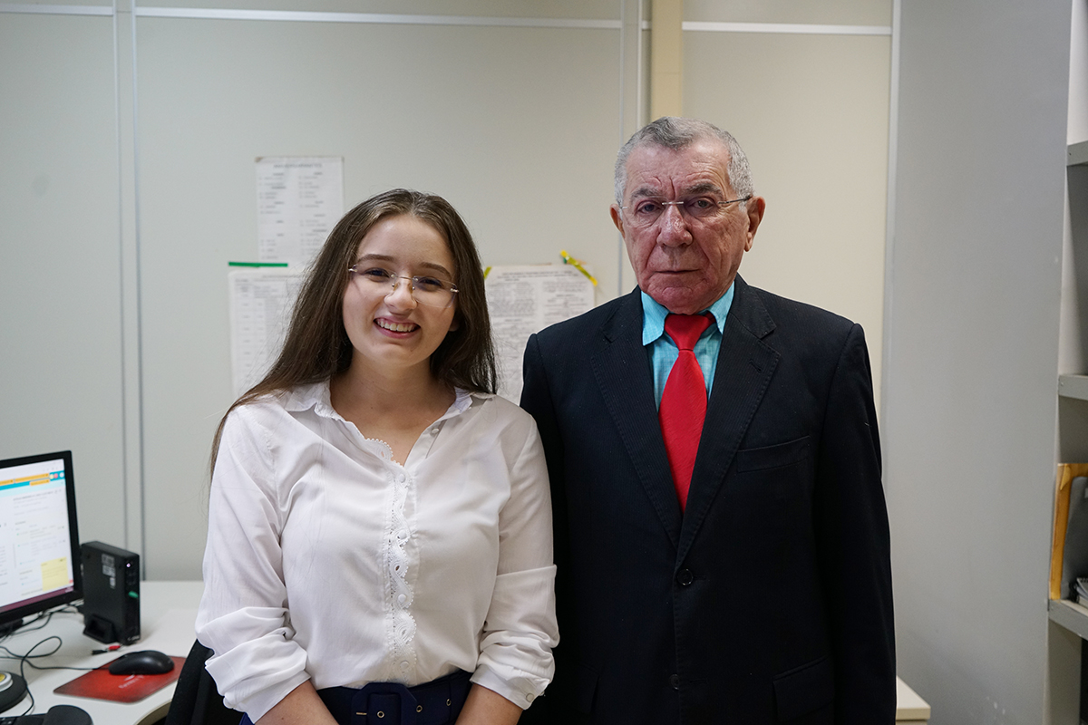 Foto da estagiária Brena e do juiz Bernardo. Estão um do lado do outro. Ela veste branco, ele paletó azul.