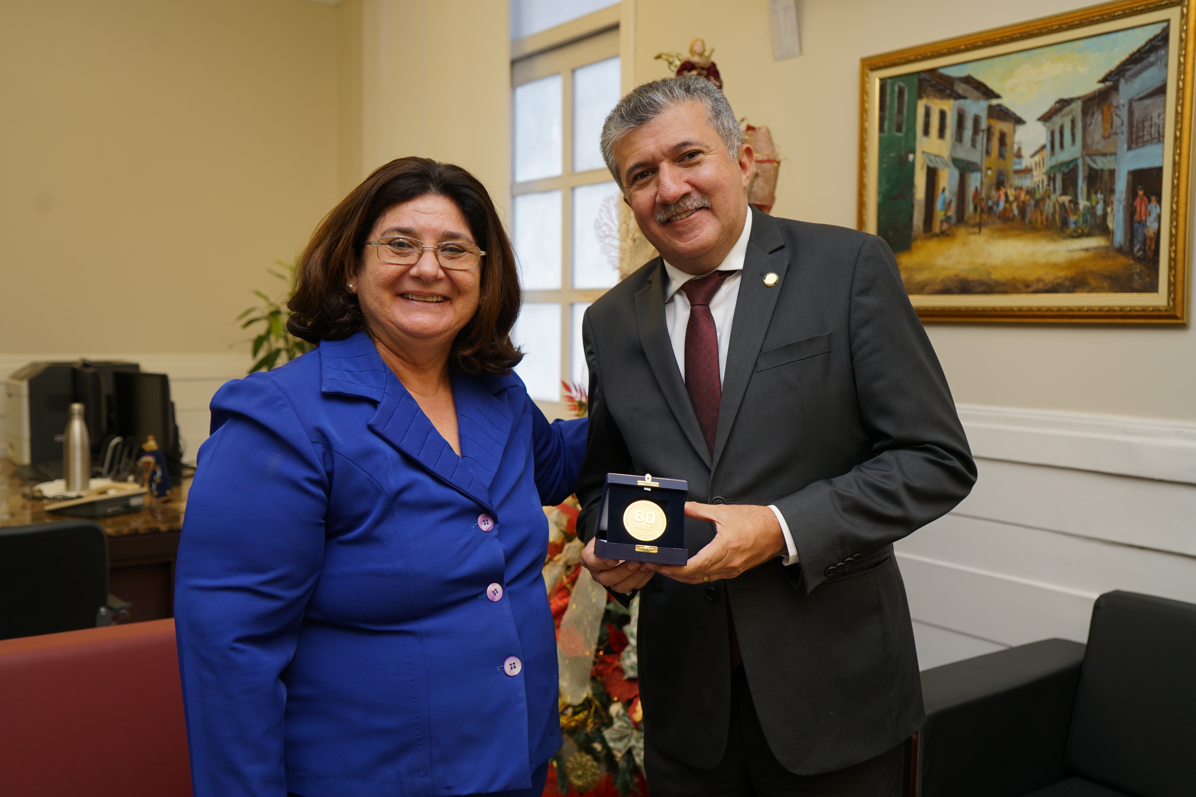 Foto do presidente da Câmara Municipal recebendo medalha comemorativa dos 80 anos da Justiça do Trabalho da Dra. Regina Gláucia observando. Ele está de paletó cinza escuro e ela, azul.