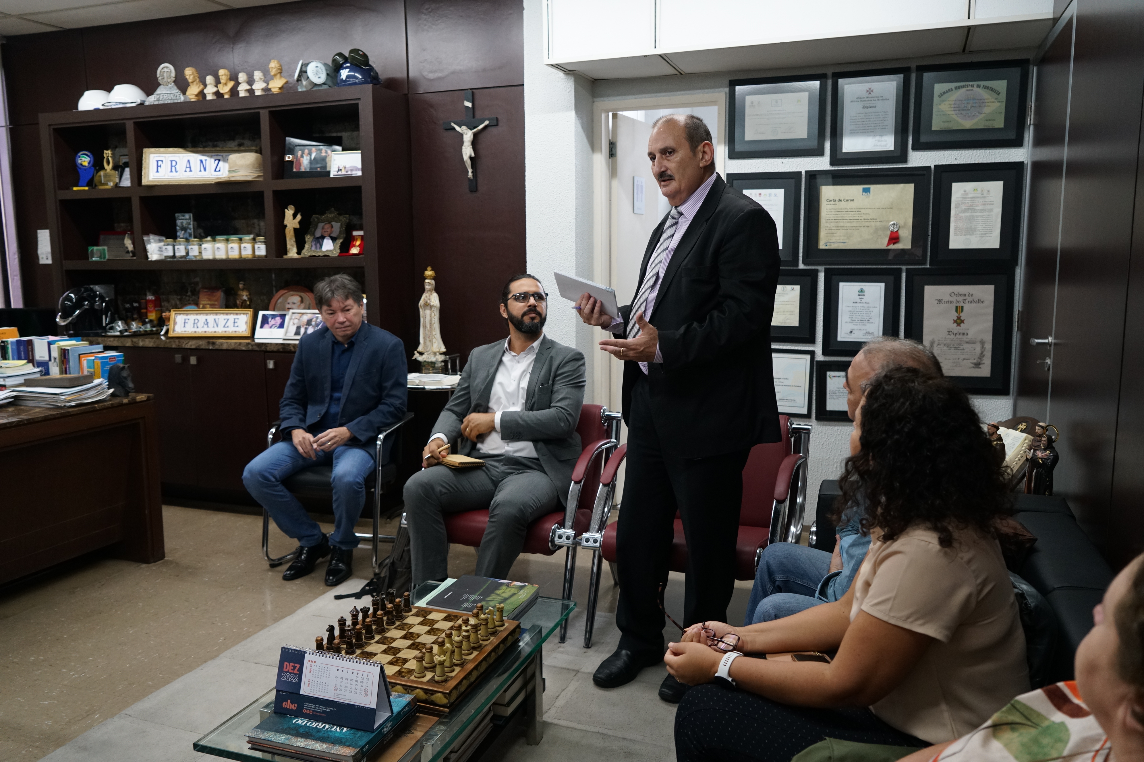 Foto em que estão alguns participantes de reunião. O des. Francisco José Gomes está em pé, no seu gabinete, falando para os presentes. Na sala, há vários objetos de arte e imagens sacras.