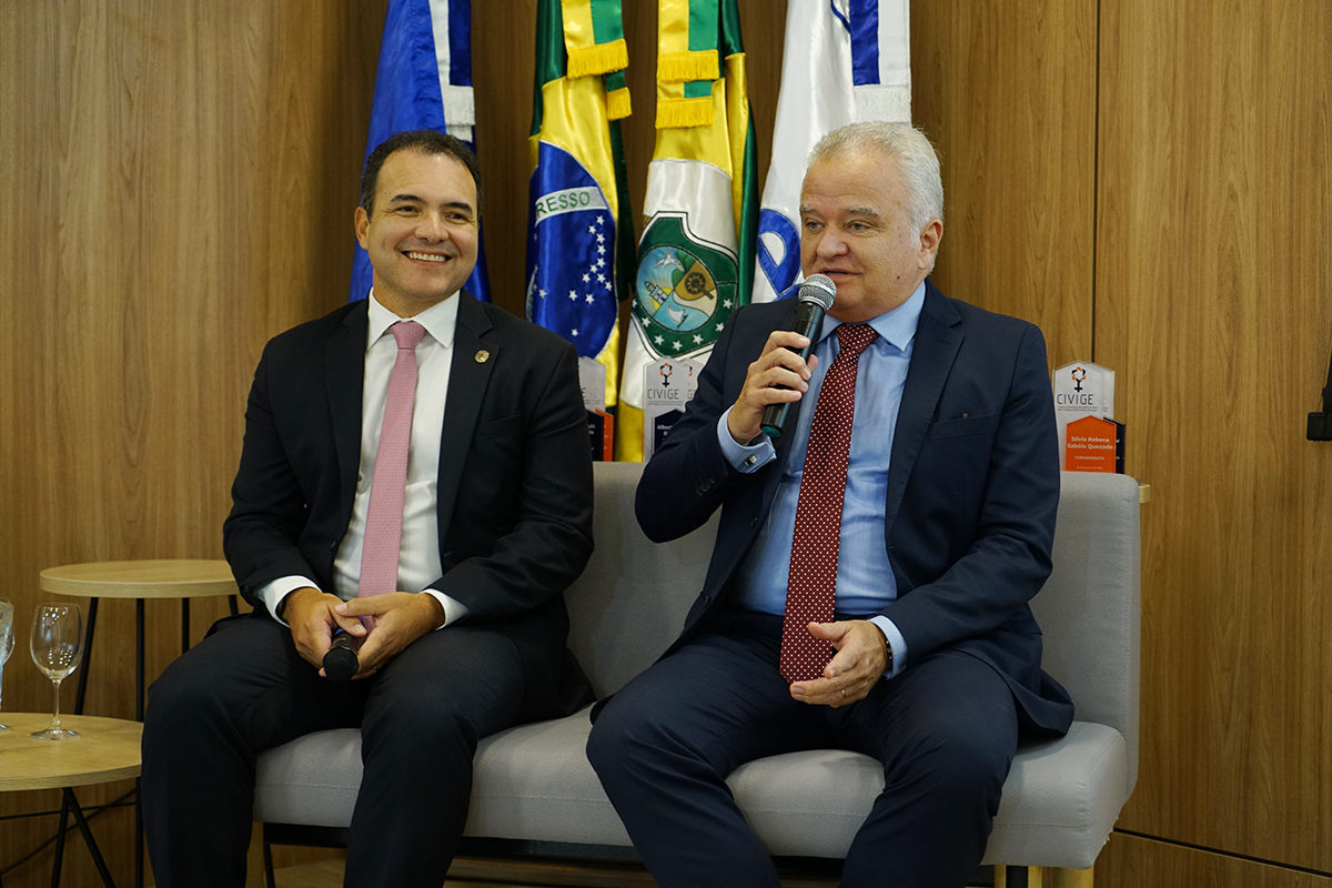 Foto do magistrado Hermano Queiroz e do conselheiro do CNJ, Marcio Luiz Coelho. Ambos estão de paletó, gravatas vermelhas e sentados num sofá. O juiz Hermano está falando ao microfone