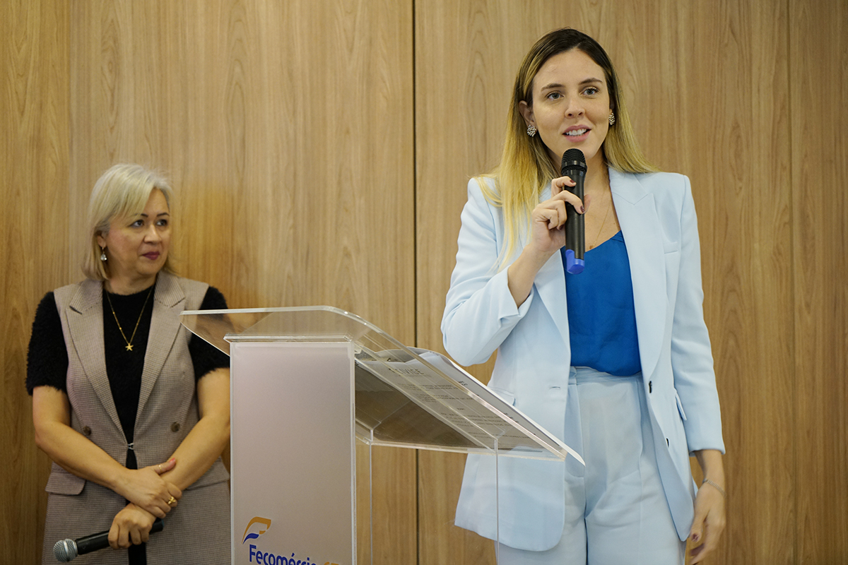 Foto da vice-governadora Jade Romero falando ao microfone em frente à tribuna. A gestora veste paletó de tom azul. Ao seu lado, consta a chefe de cerimonial que está de roupa cáqui e preta. Ambas estão sorridentes