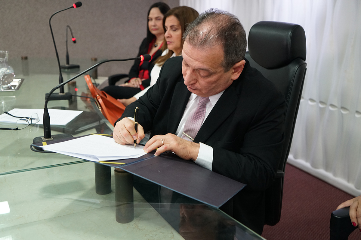 Foto do Dr. Durval Maia assinando o documento. Ele usa paletó preto e gravata rosa claro
