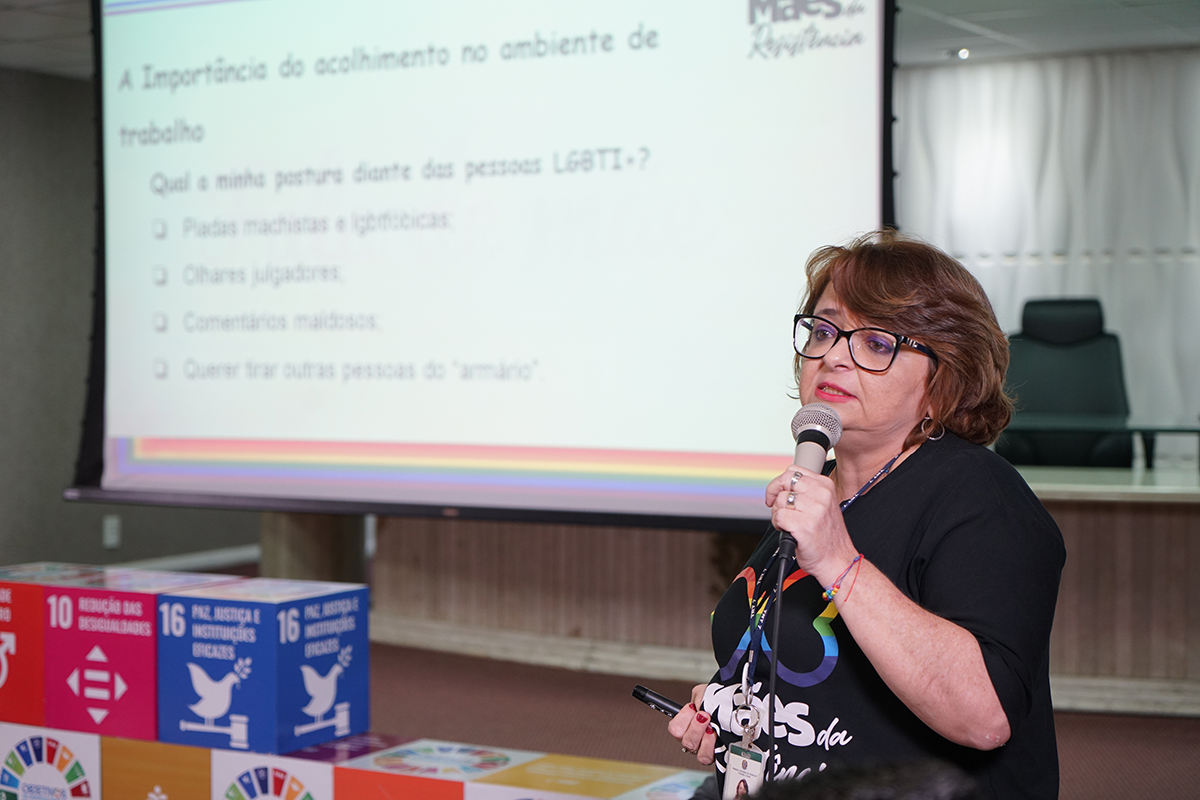 Foto de Cristina segurando um microfone. Ela usa óculos e uma blusa preta com a frase Mães da Resistência