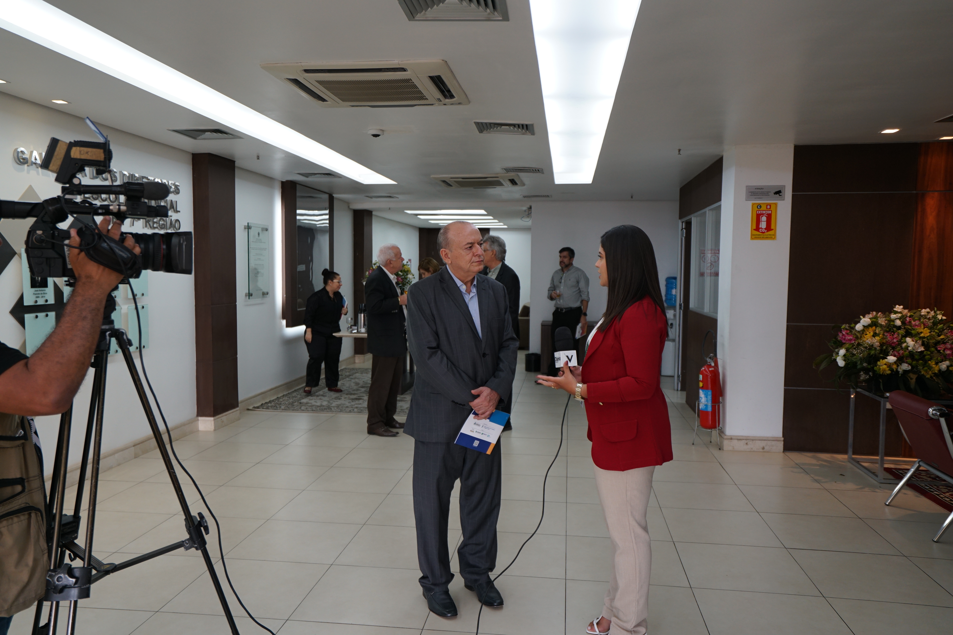 Foto do des. José Antônio Parente sendo entrevistado pela repórter. Ele está de paletó cinza e ela de blazer vermelho. Na foto, consta ainda a câmera que está filmando a entrevista. Há algumas pessoas atrás deles, no momento do coffee break