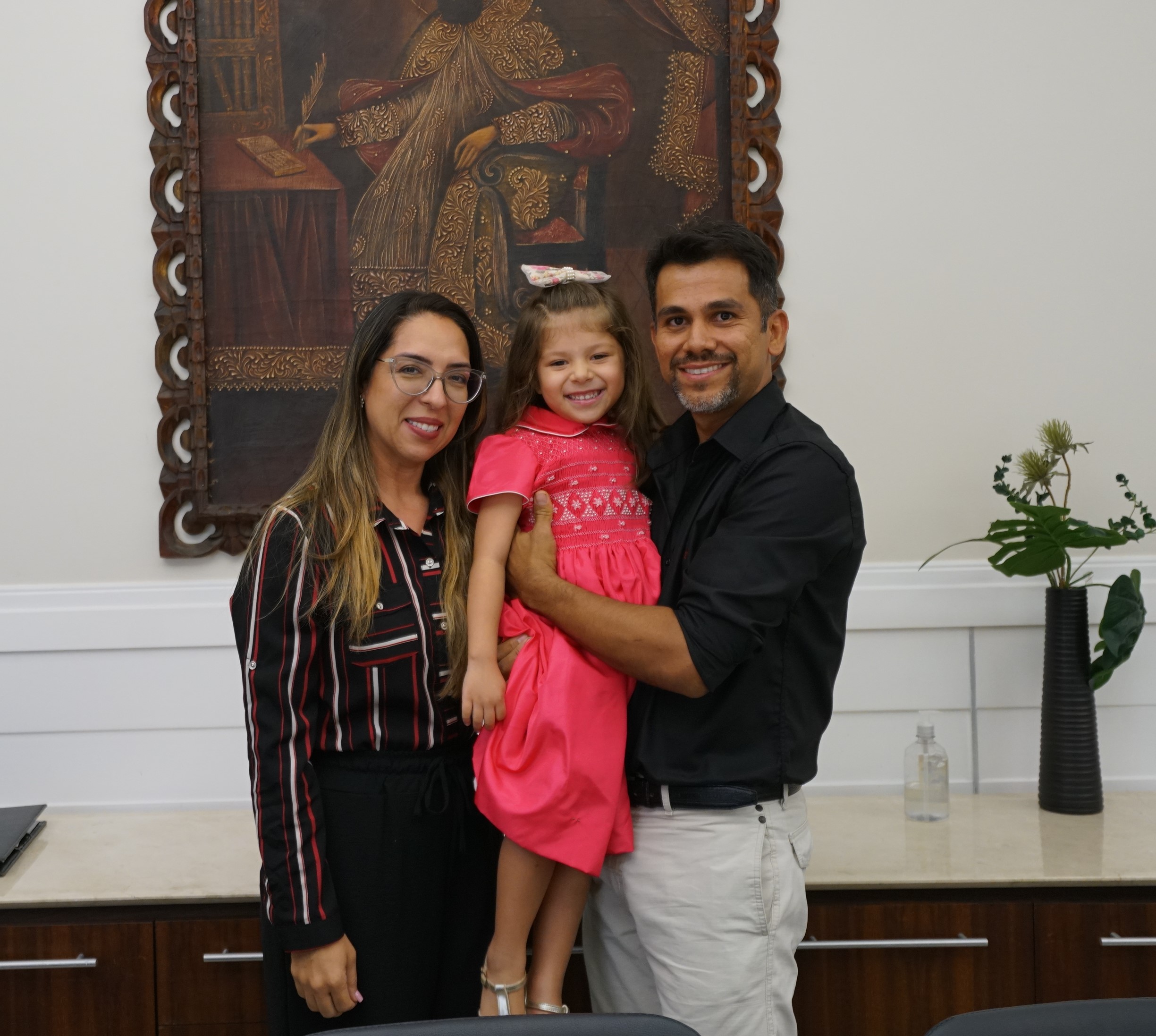 Foto na Presidência da posse da servidora Ana Paula acompanhada da filha e do esposo. Eles estão sorridentes e existe uma obra de arte (quadro) no fundo da foto.