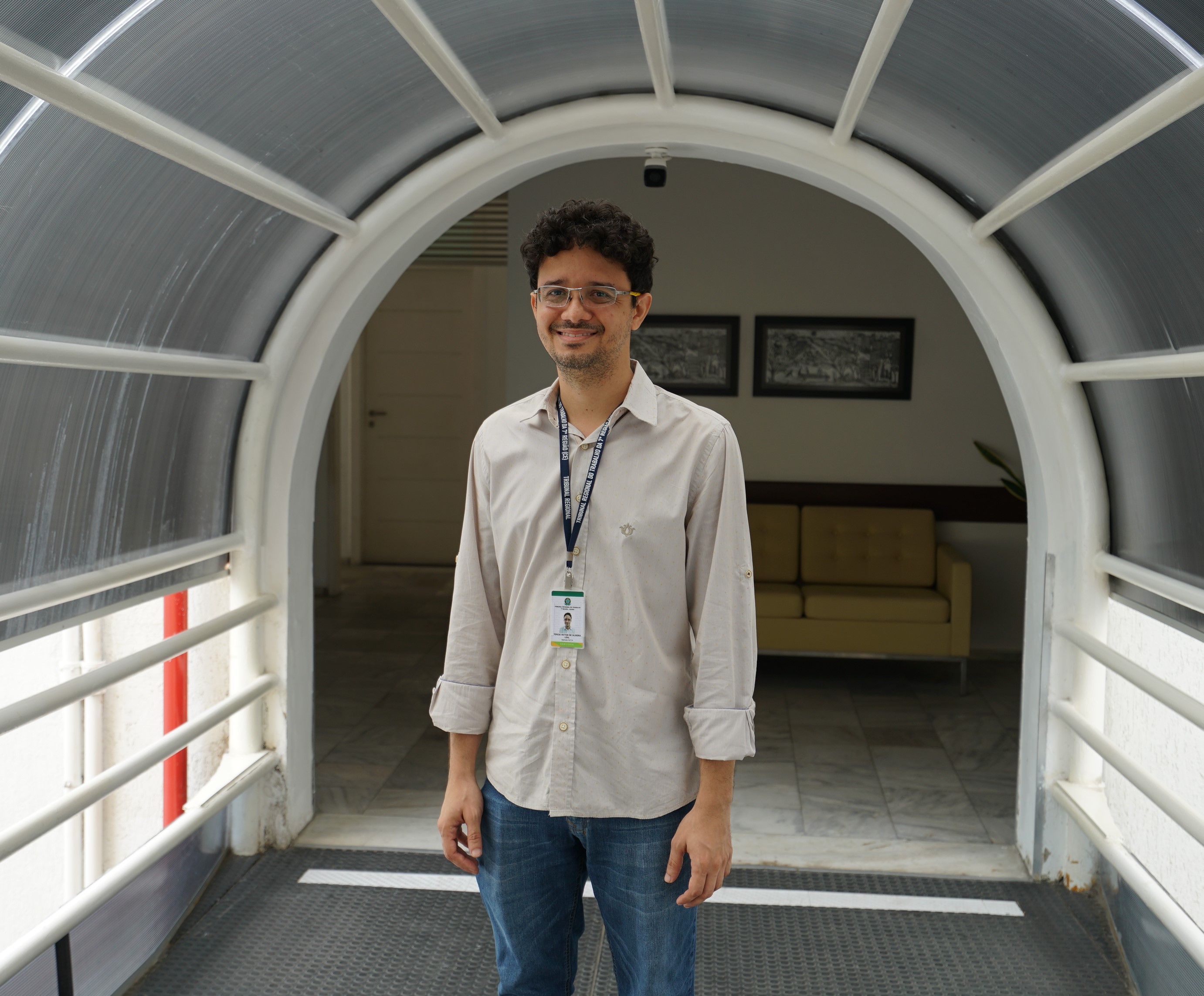 Foto de Tércio sorrindo num corredor do anexo II do TRT-7. Ele está sorridente, usa óculos e crachá. Está vestindo uma camisa social de cor clara.