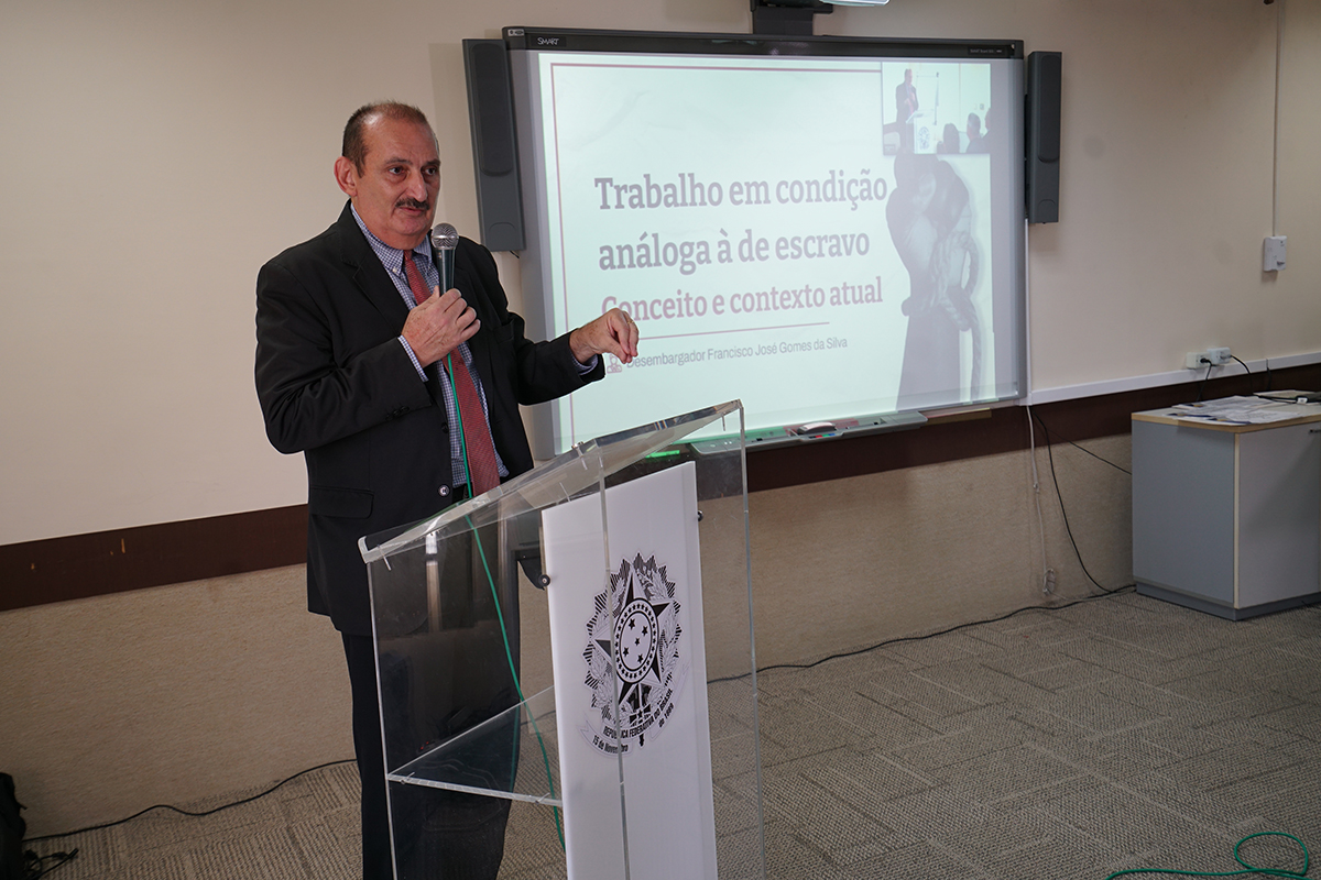 Homem de terno fala ao microfone com slide atrás