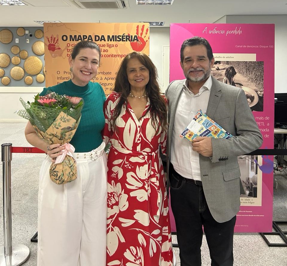 Foto das Juízas do trabalho Kaline Lewinter e Ivânia Araújo e o magistrado Ronaldo Solano. Kaline está segurando um buquê de rosas, Ronaldo segura um presente e eles estão sorridentes e abraçados à diretora do Fórum, Ivânia Araújo. Atrás deles estão vários cartazes de campanhas de programas da Justiça do Trabalho