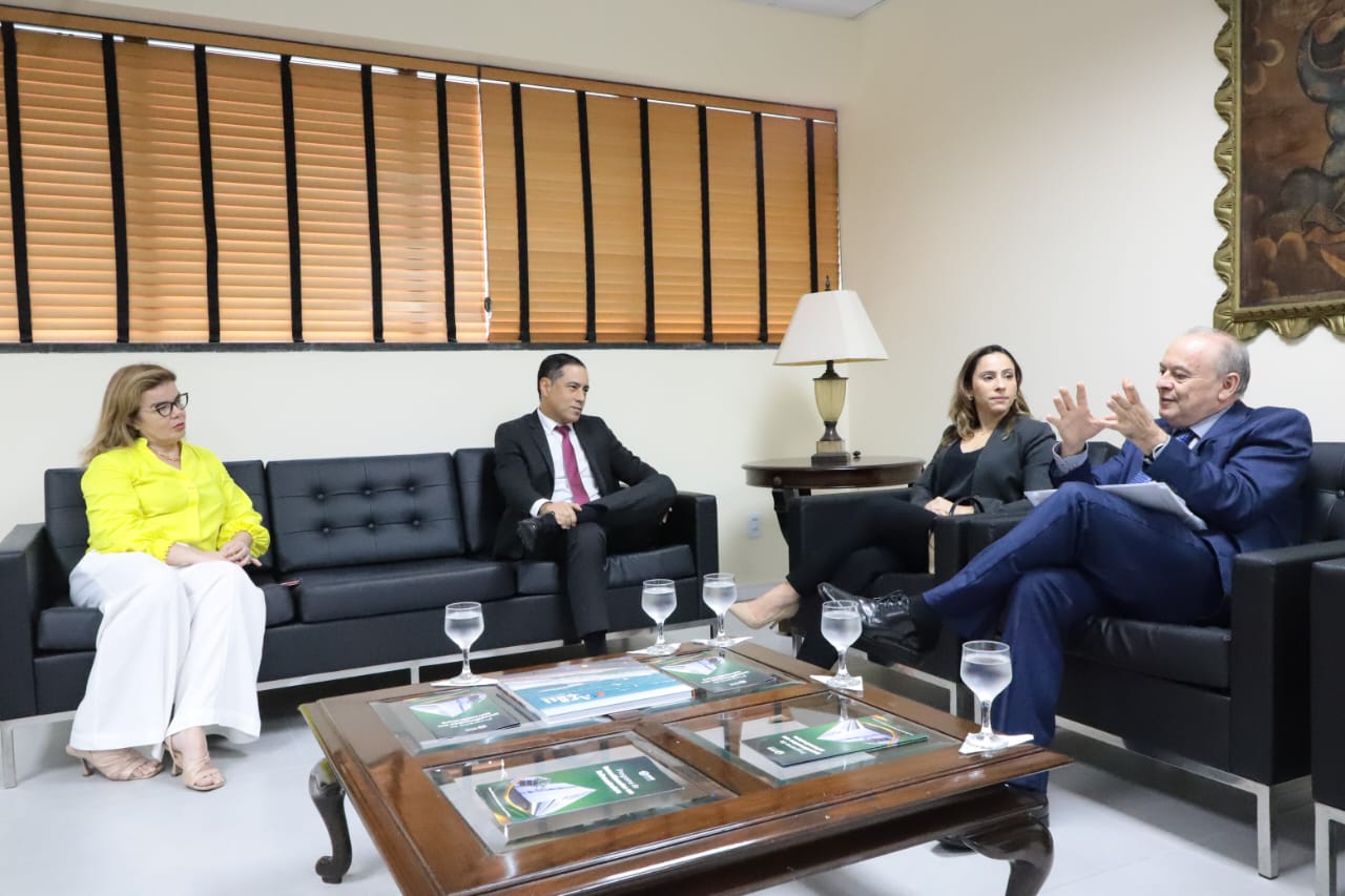 Foto na sede da PGJ em que os participantes (4 pessoas) estão sentados em sofás pretos. O des. Parente está falando e gesticulando, enquanto o procurador geral de justiça, a assessora de Desenvolvimento Institucional, promotora de Justiça Daniele Fontenele e a juíza do Trabalho substituta, Naira Alencar, estão olhando para ele. Na sala onde ocorre a reunião há persianas, uma pintura, um abajour e uma mesa de madeira com copos de água