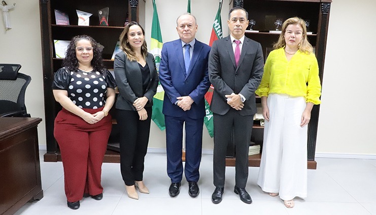 Foto dos 5 participantes da reunião em pé. São três mulheres e dois homens. Na parede atrás da foto, constam três bandeiras e estantes