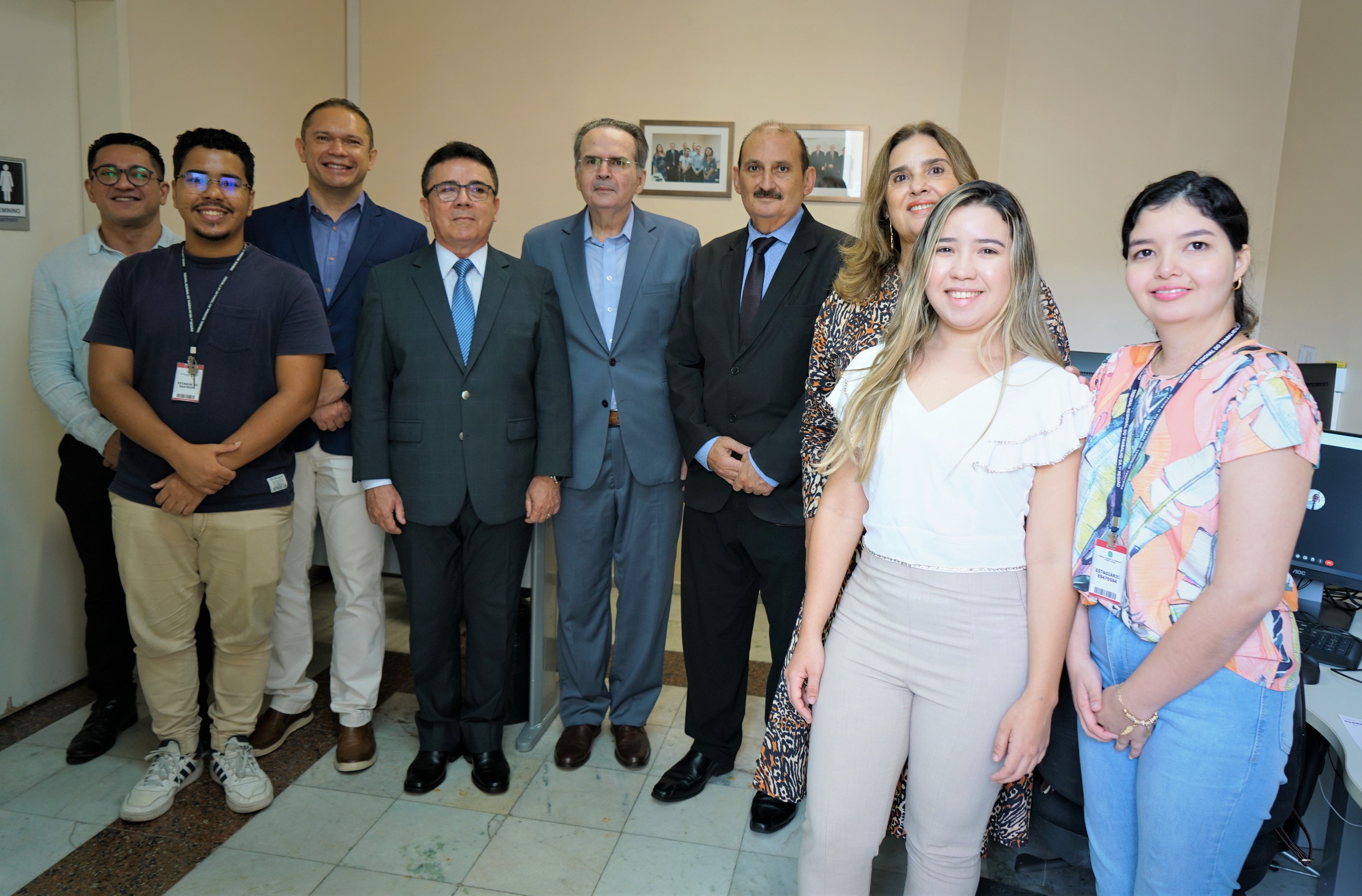 Foto da equipe da 2ª turma. Eles estão em pé, sorridentes. Os três desembargadores e o secretário da turma vestem paletó. Alguns estagiários de crachá. Atrás deles, estão fixados os dois retratos na parede, que é bege.