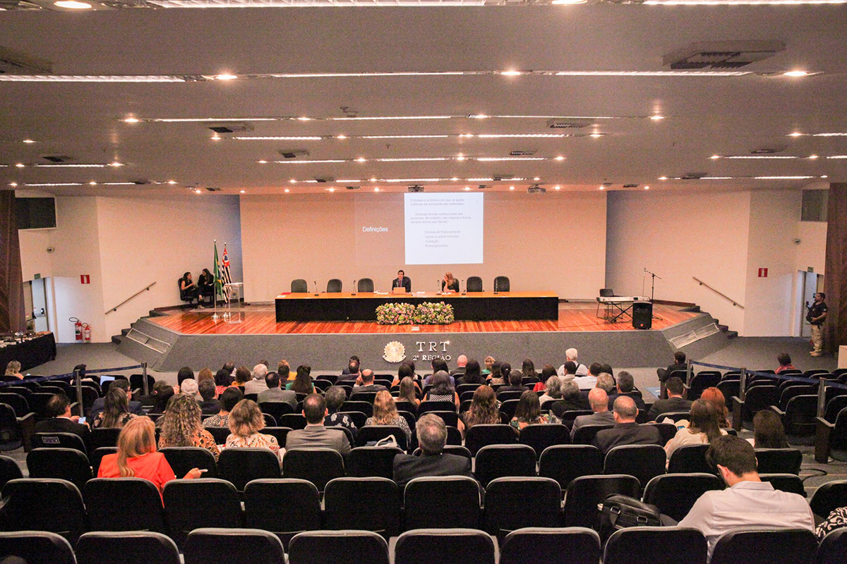 Grande auditório com dezenas de pessoas sentadas de frente para uma mesa e um painel