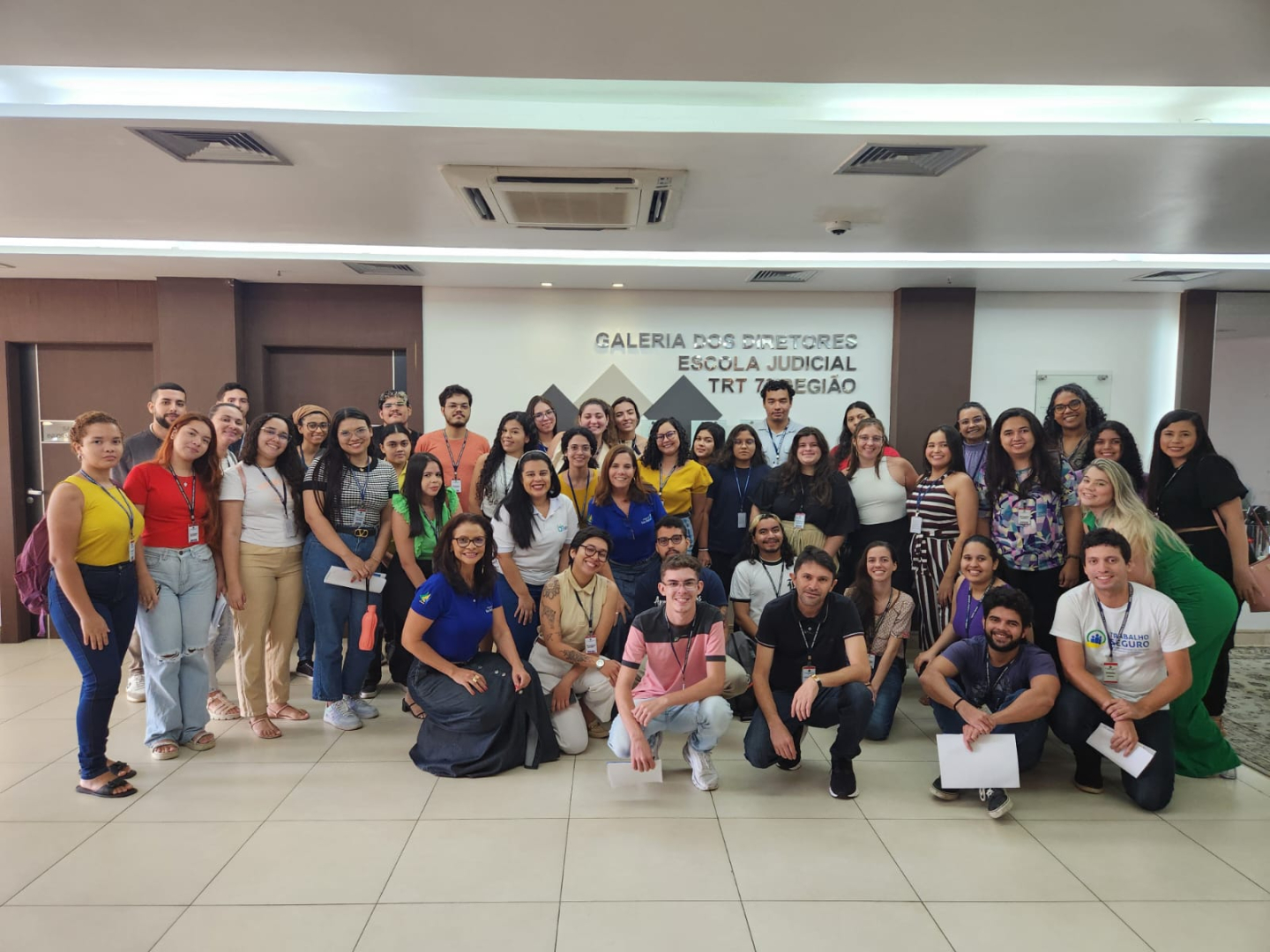 Foto do grupo de estagiários(as) junto com duas integrantes do Programa Fada Madrinha e a expositora do Sine, Carliane. Eles estão sorridentes, alguns em pé, outros agachados e estão no hall da Escola Judicial. Há em torno de 25 a 30 pessoas na imagem