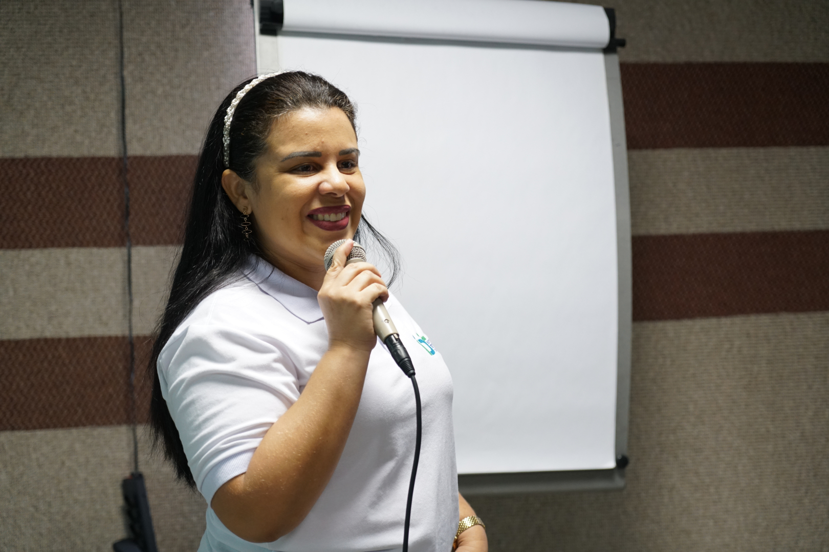 Foto da Psicóloga do IDT / Sine, Carliane Anastácio. Ela está segurando um microfone, sorridente, usa blusa branca com a logomarca do Sine.