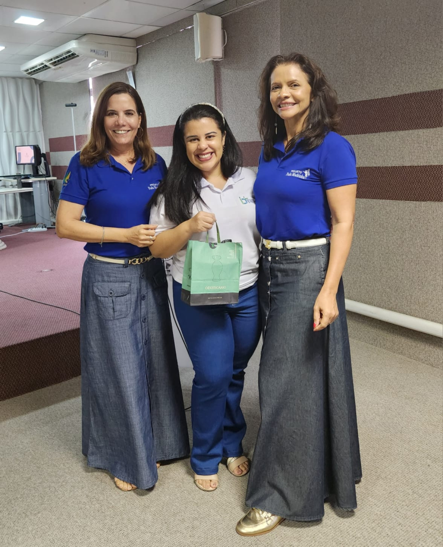 Foto das Integrantes do subcomitê Neuza Sucupira e Carmem Moura. Elas estão entregando lembrança à psicóloga do Sine, sorridentes e vestem a blusa do programa Fada Madrinha na cor azul com a logo do programa