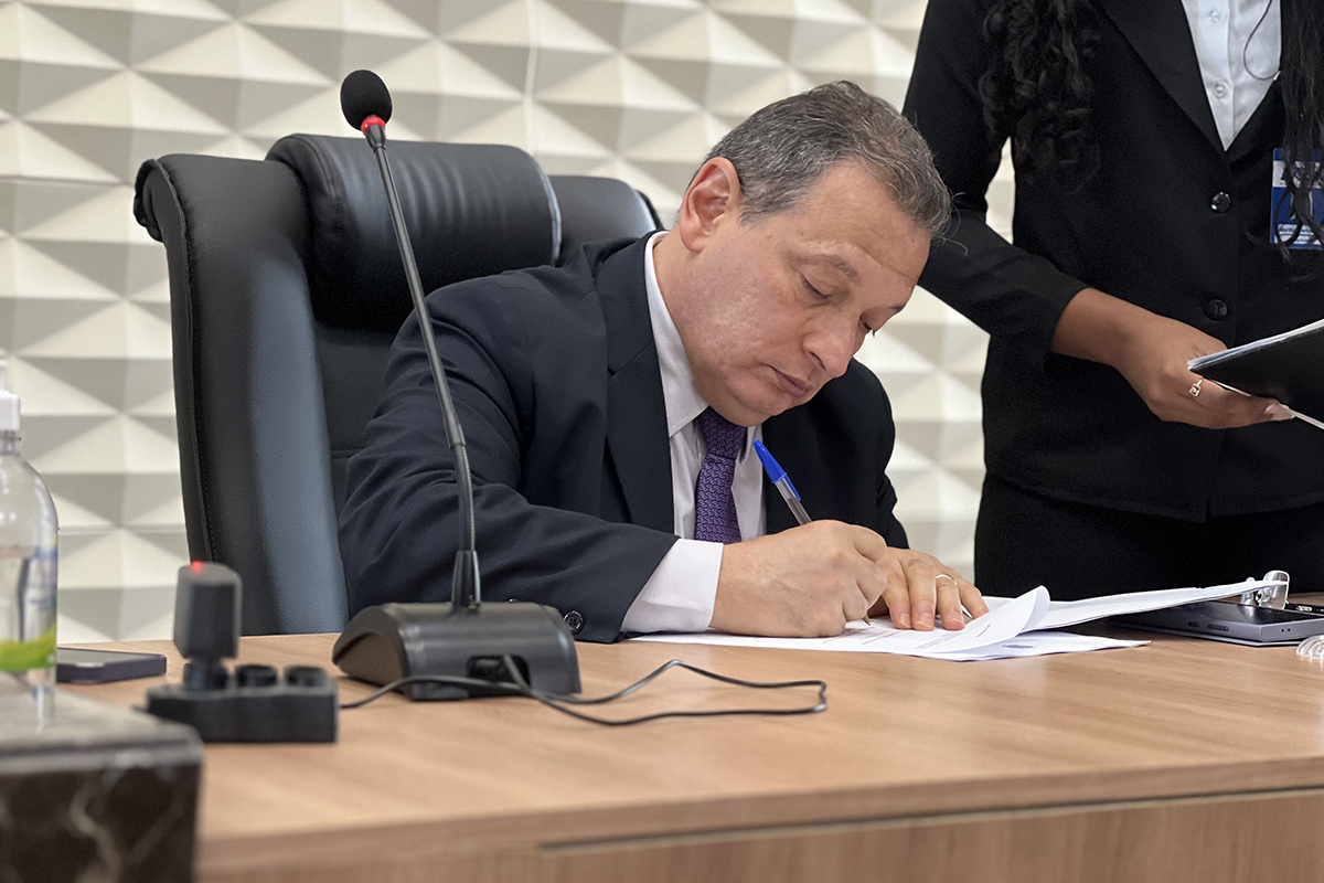 homem de paletó assina um papel com uma caneta bic azul em uma mesa com um microfone