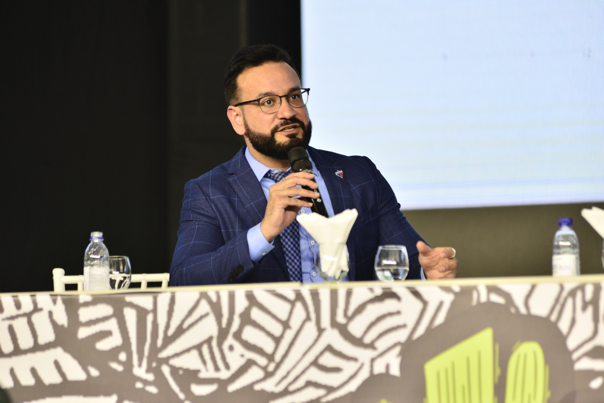 homem de paletó fala ao microfone na mesa do evento