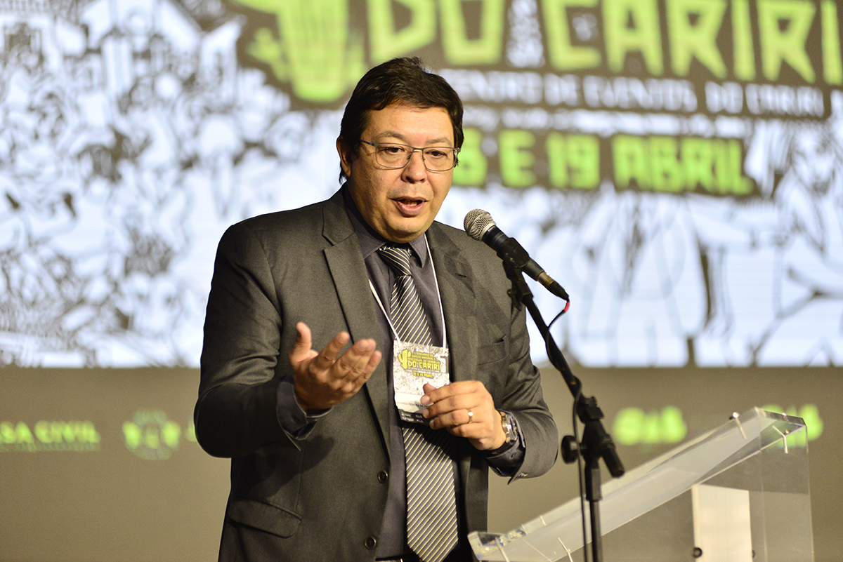 homem de paletó fala ao microfone na tribuna do evento com painel atrás