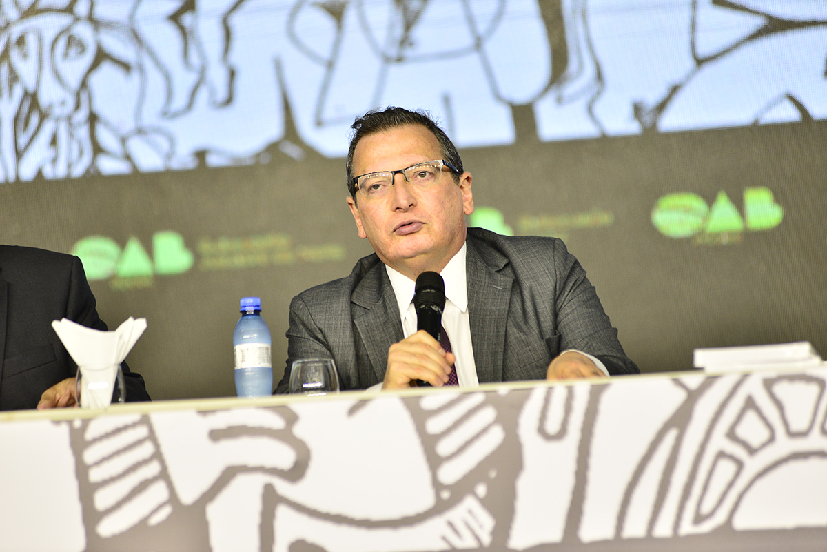 homem de paletó fala ao microfone na mesa do evento com painel atrás