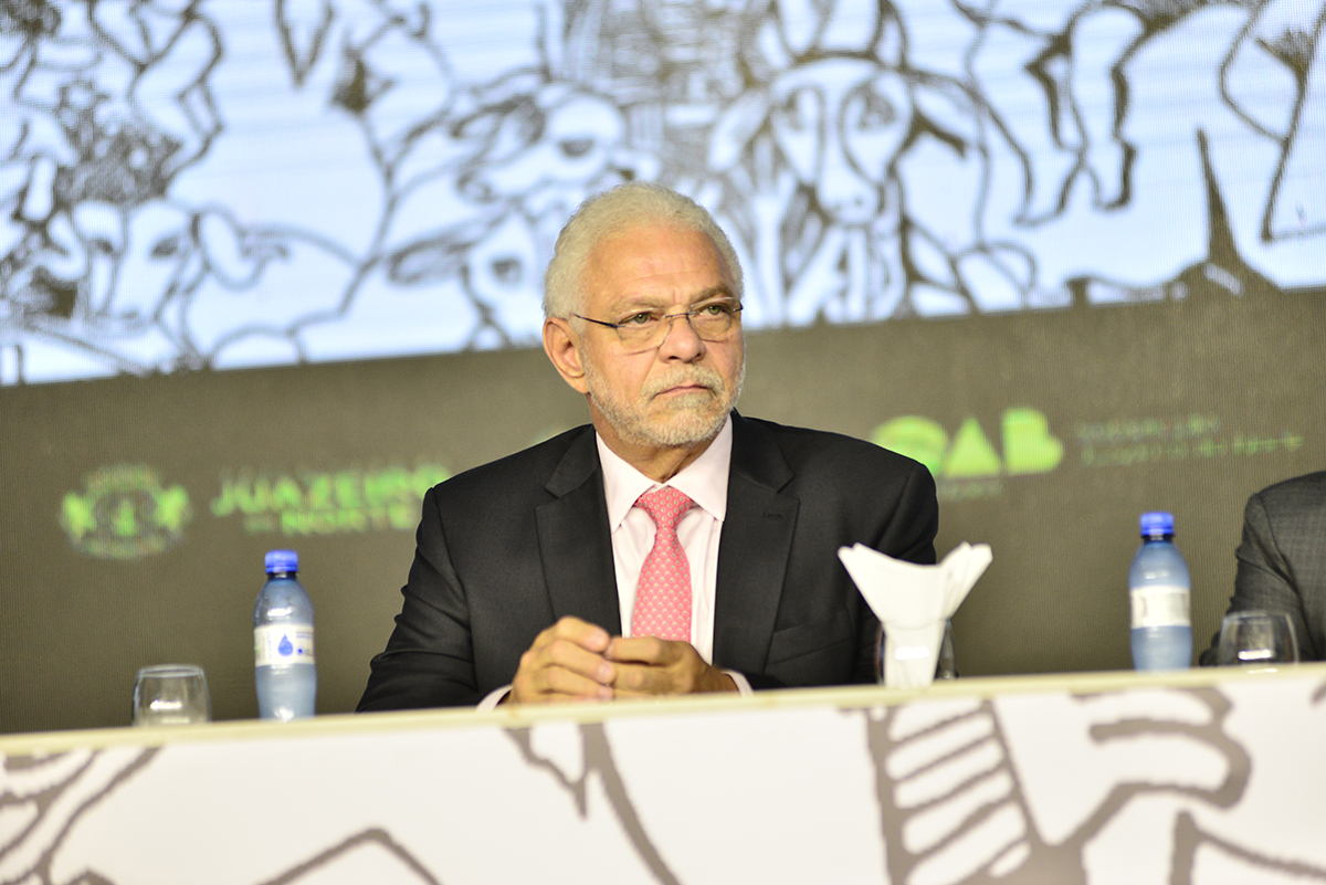 Homem de paletó na mesa do evento com painel atrás