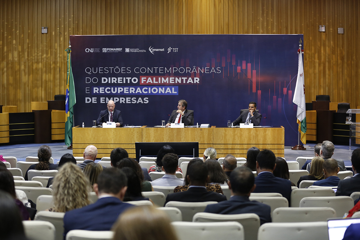 mesa com três palestrantes, em um auditório de paredes de taco de madeira