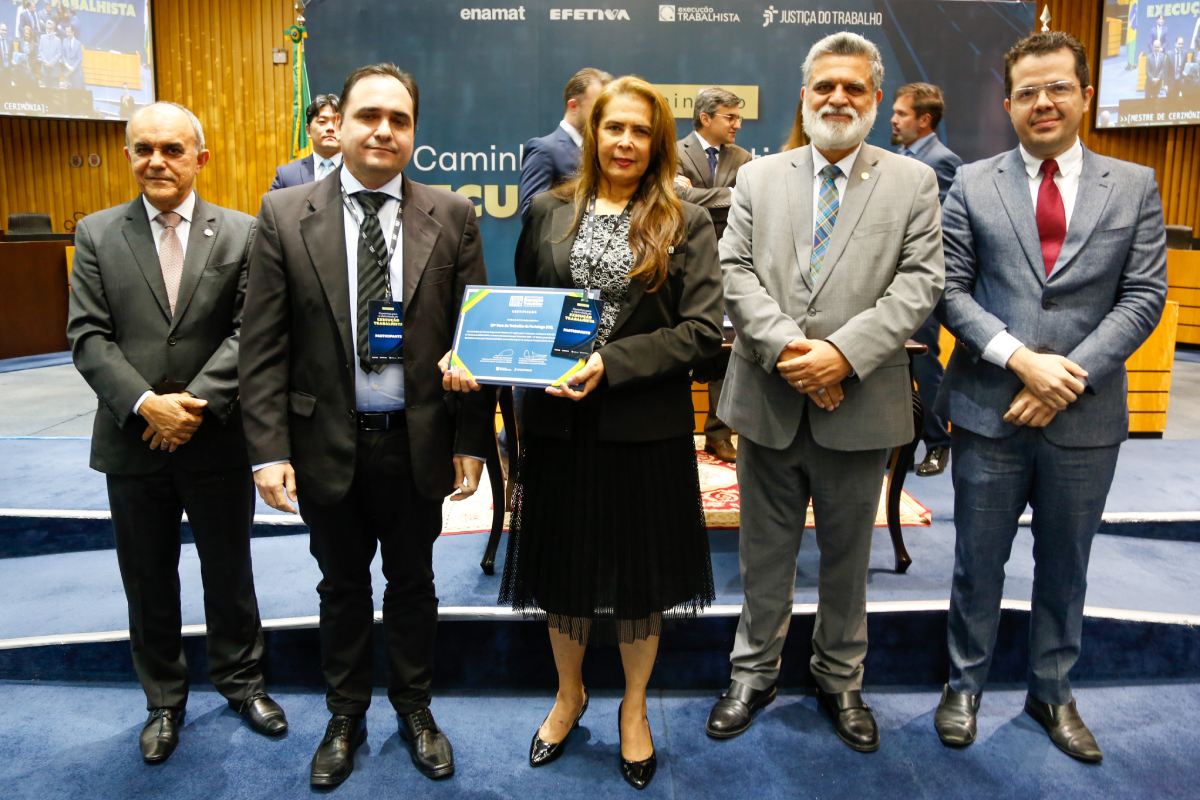 quatro homens de paletó em pé, uma mulher em pé segurando um certificado, num auditório de taco de madeira nas paredes e carpete azul