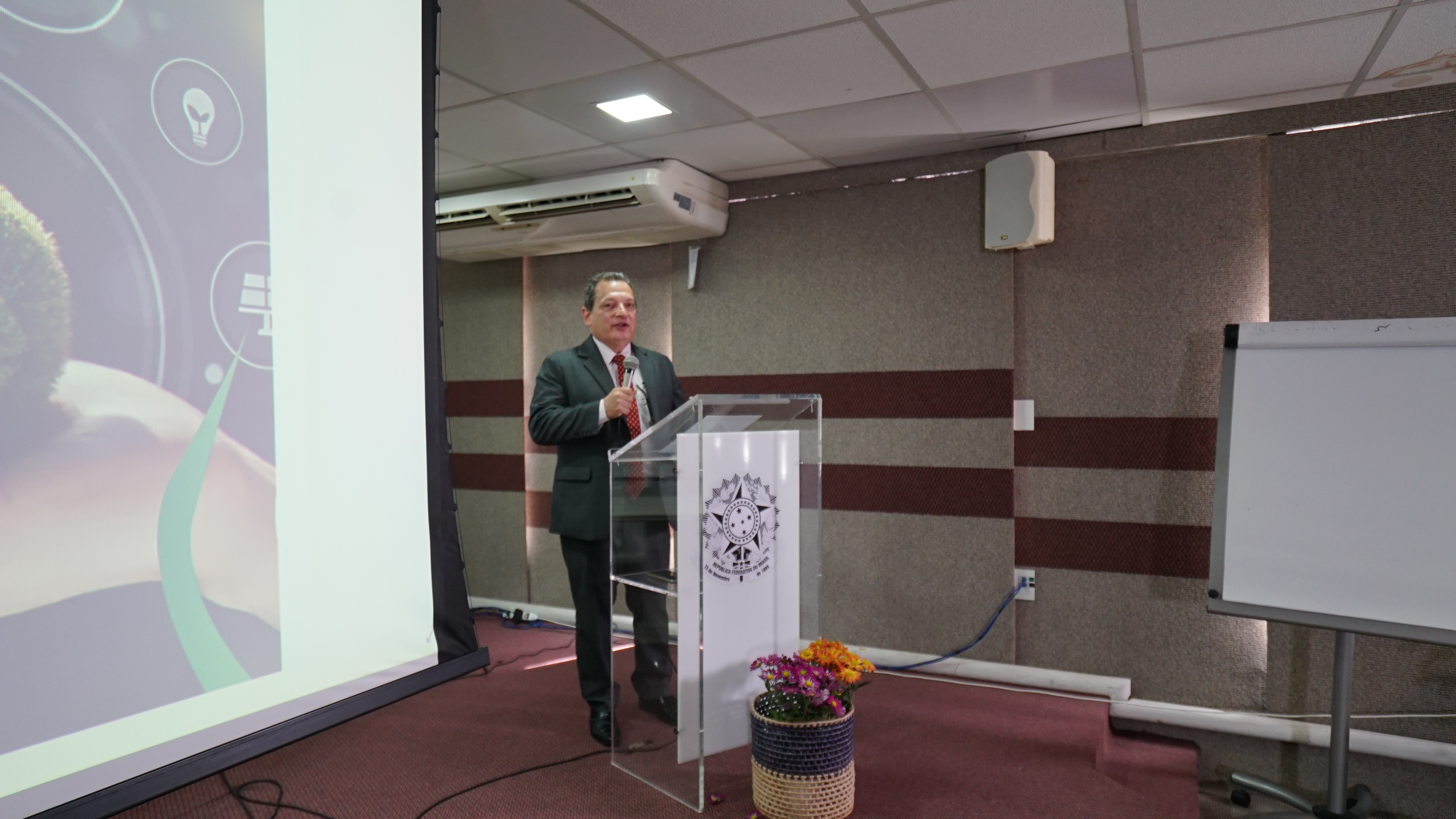 Foto do presidente Durval César falando na tribuna do auditório. Ele está segurando um microfone e veste um paletó cinza e gravata vermelha. Em frente à tribuna de acrílico, há um cesto de palha com rosas naturais