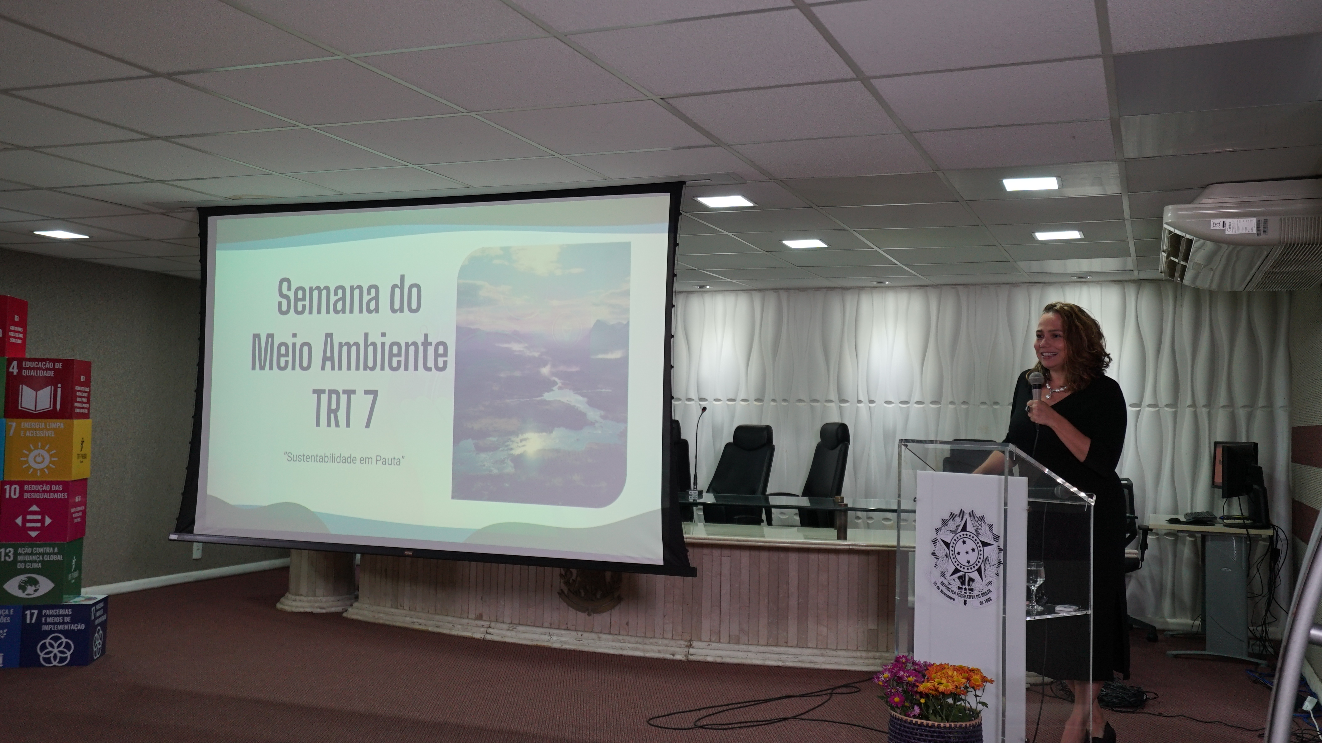 Foto da palestrante Victoriana Gonzaga falando na tribuna de acrílico com a logomarca do TRT7. Ela segura um microfone, está usando um vestido e sandálias pretas e está sorrindo no momento que está falando. Ao seu lado, tem uma tela com a projeção de slide que está escrito: "Semana do Meio Ambiente TRT7"