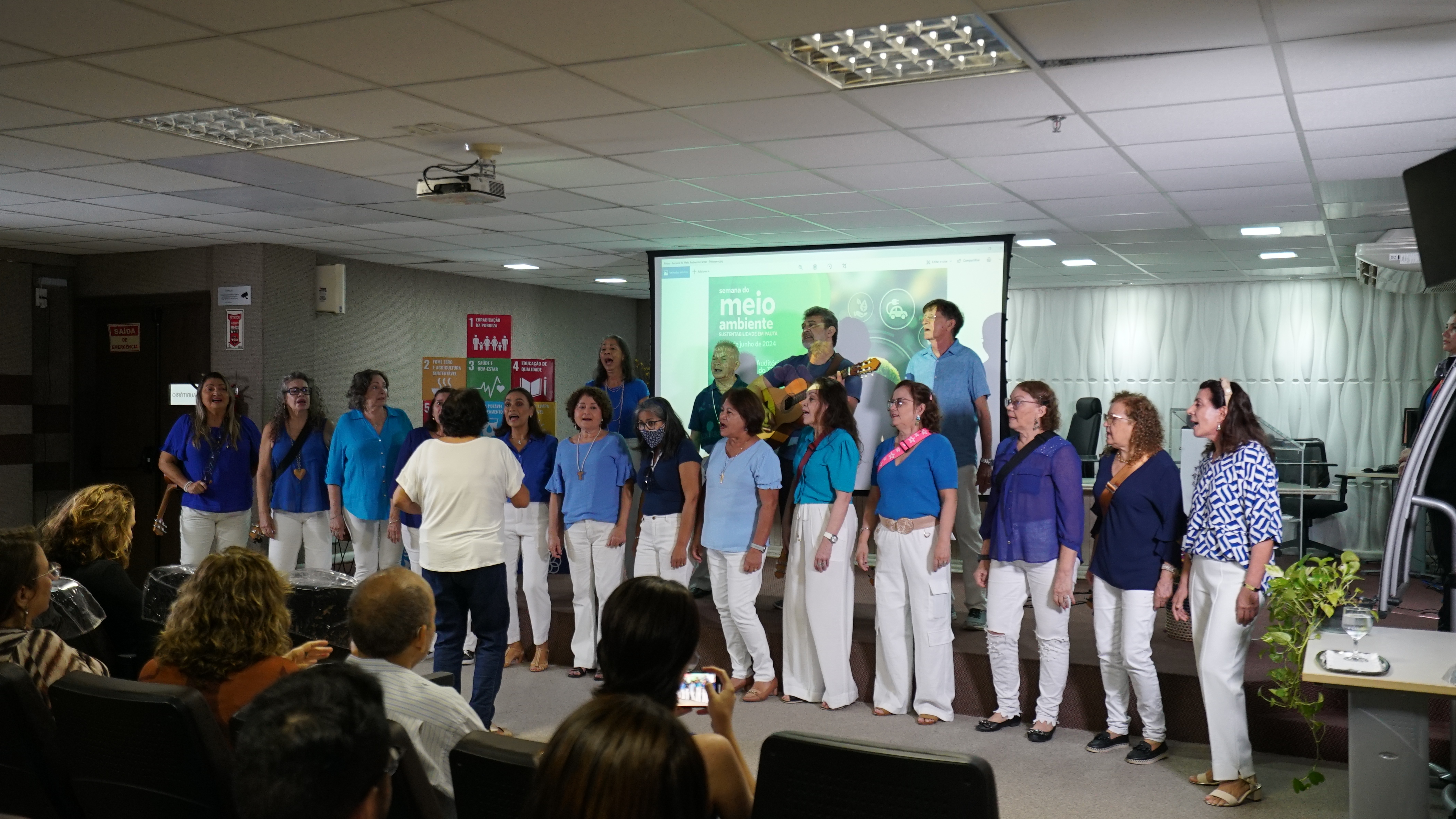 Foto do coral Sétima Voz cantando no auditório em cima do palco. Homens e mulheres estão vestindo blusas de cor azul e calças brancas. As pessoas no auditório estão assistindo à apresentação