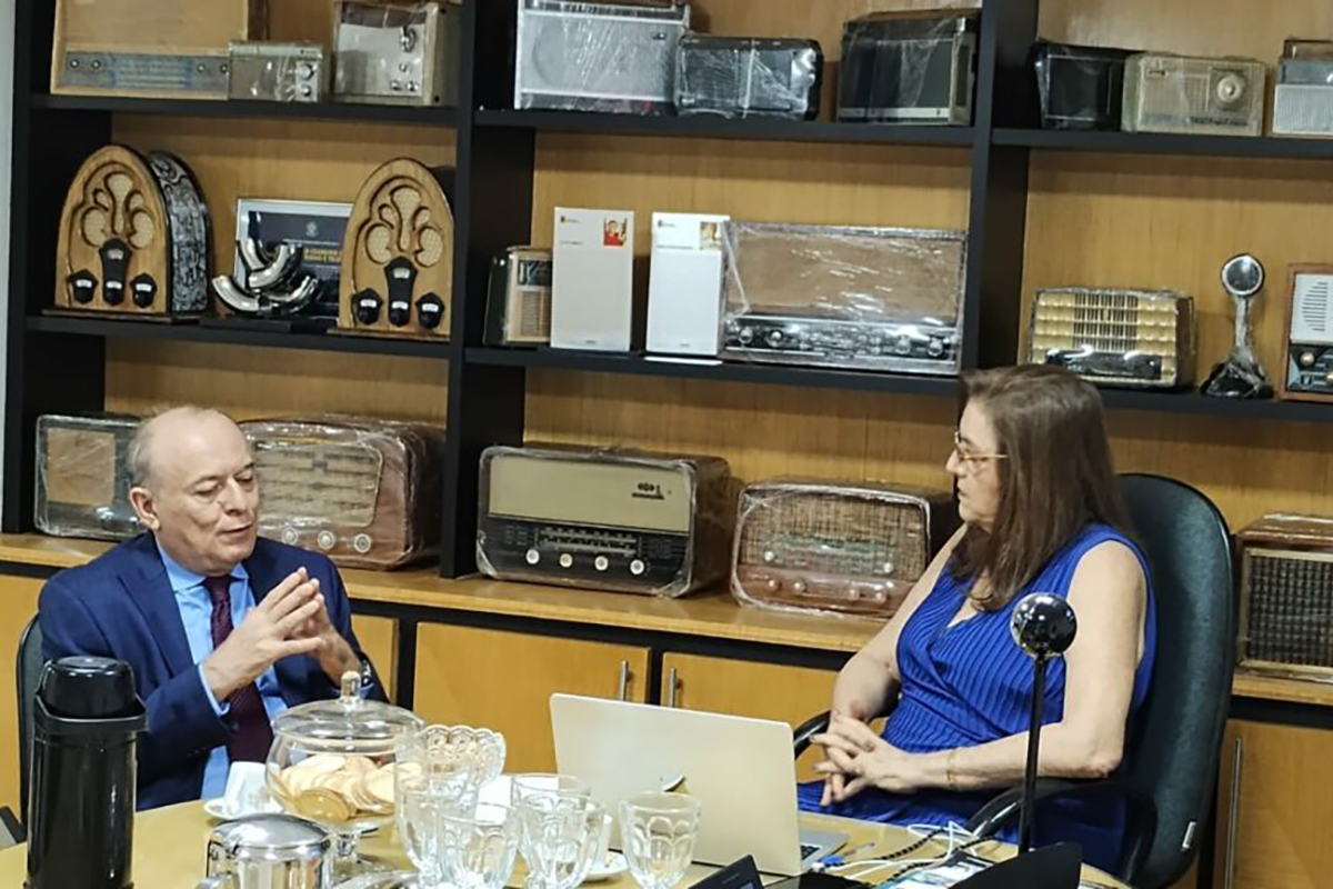 Homem de terno conversa com mulher de vestido azul numa mesa. Atrás deles, uma grande estante exibe aparelhos de rádio e televisão antigos