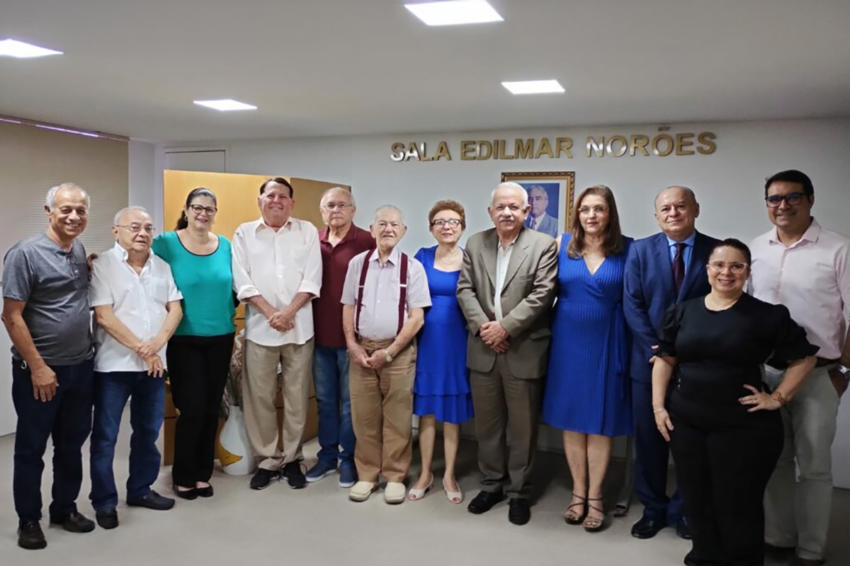 Doze pessoas em pé posam para foto numa sala de piso e parede brancos, ao fundo o retrato de um homem e o letreiro Sala Edilmar Norões