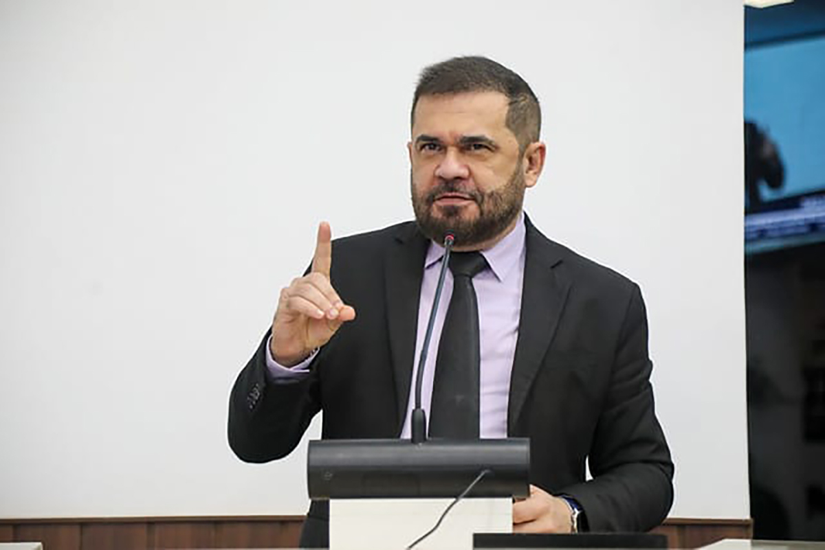 homem de paletó e gravata fala ao microfone com um dedo levantado