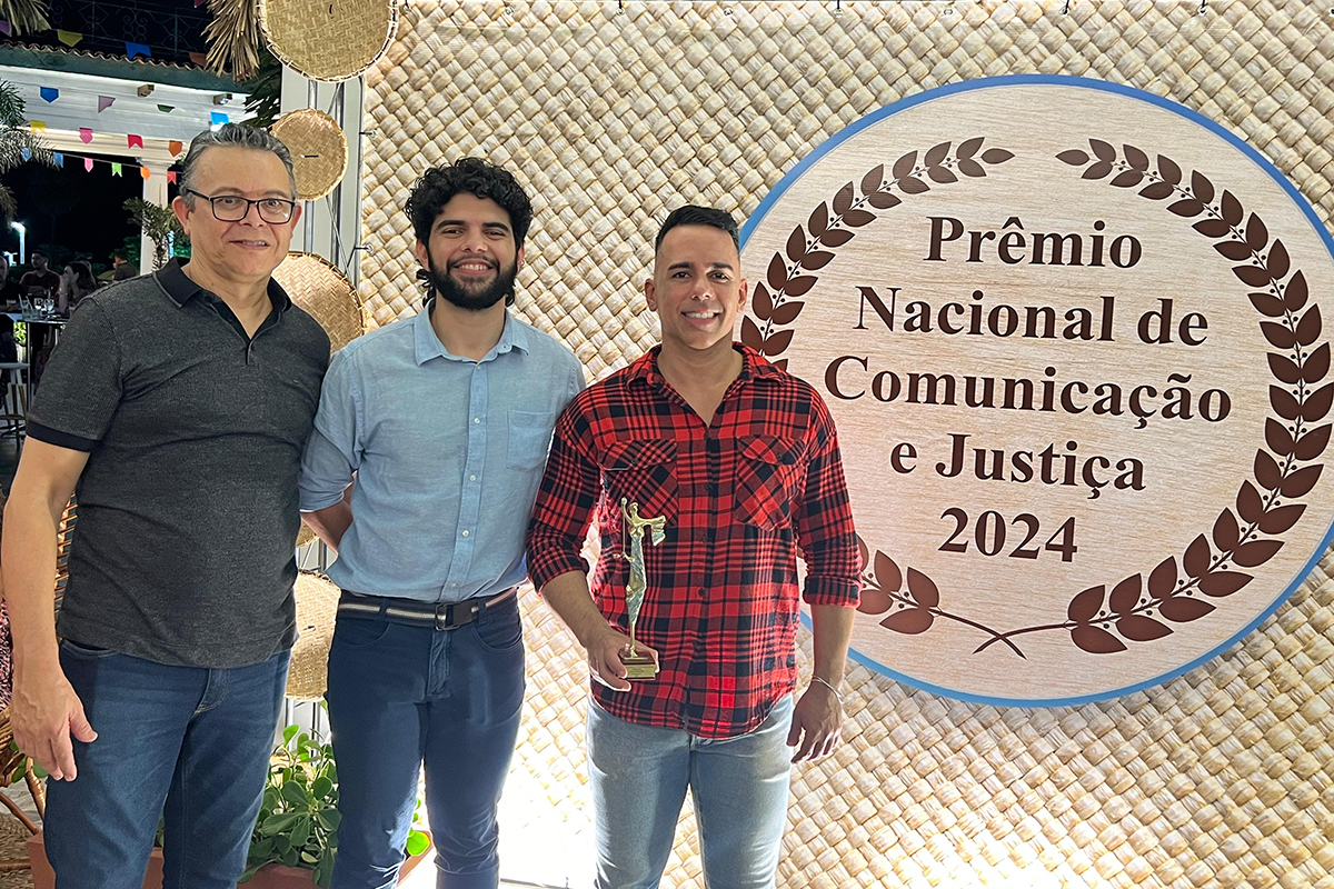 Três homens em pé na frente de um painel com decoração junina e o nome pintado: Prêmio Nacional de Comunicação e Justiça 2024. O homem da direita segura um troféu, que é uma estátua da deusa da justiça
