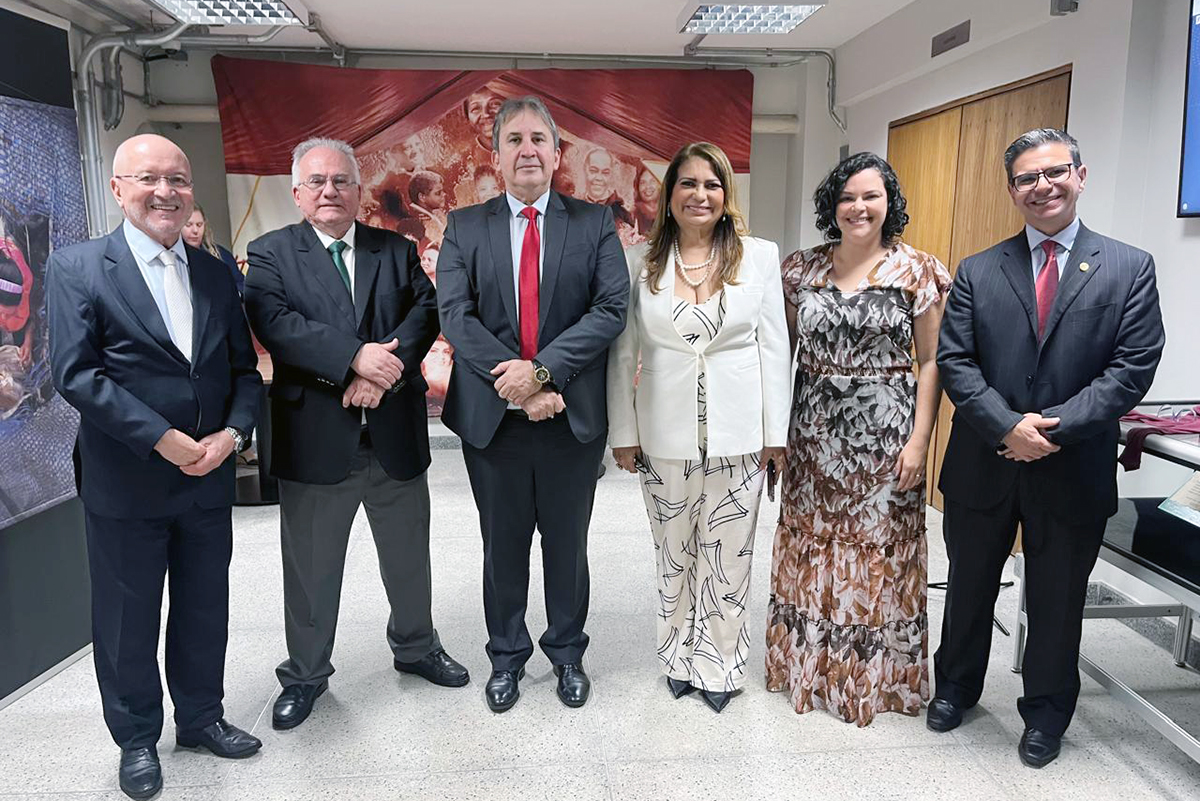 Quatro homens em pé de terno e gravata, duas mulheres, uma de blazer e outra de vestido. Eles estão numa sala ampla, com paineis nas paredes e uma porta de madeira do lado direito.