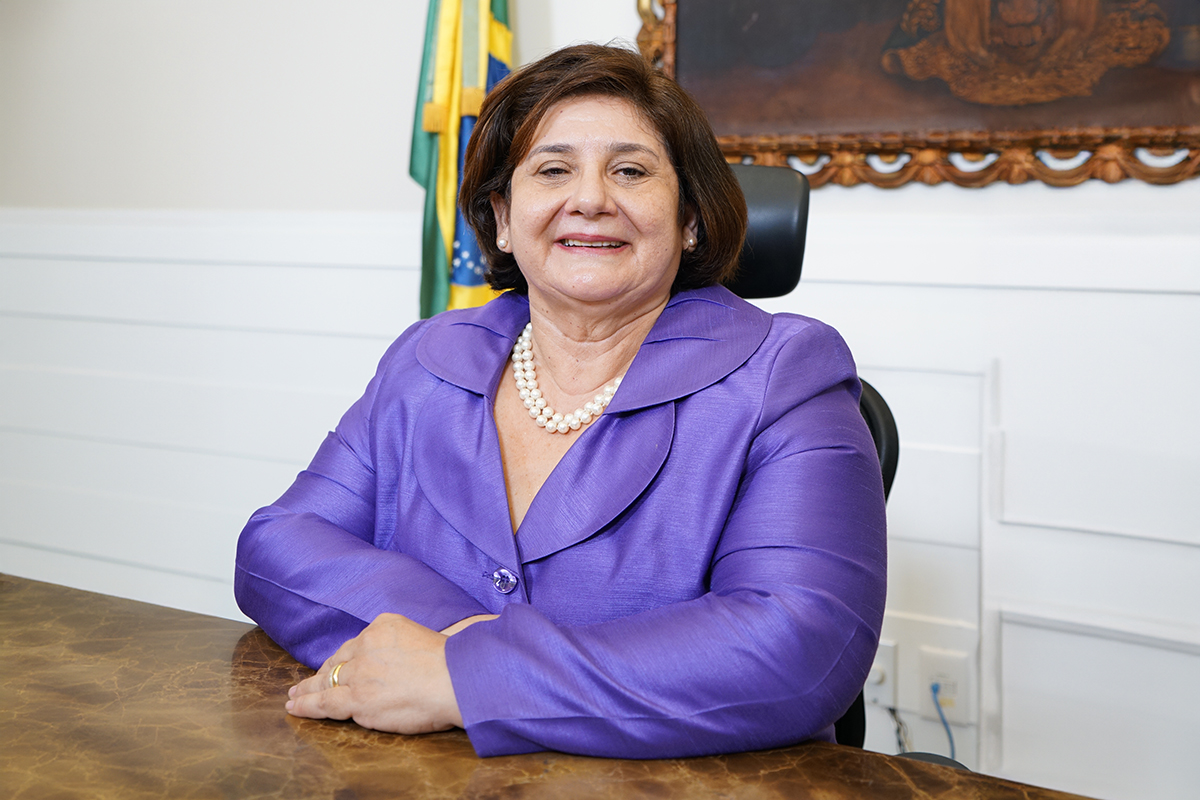 Foto da desa. Regina Gláucia. Ela veste blazer lilás, está sentada e sorridente.