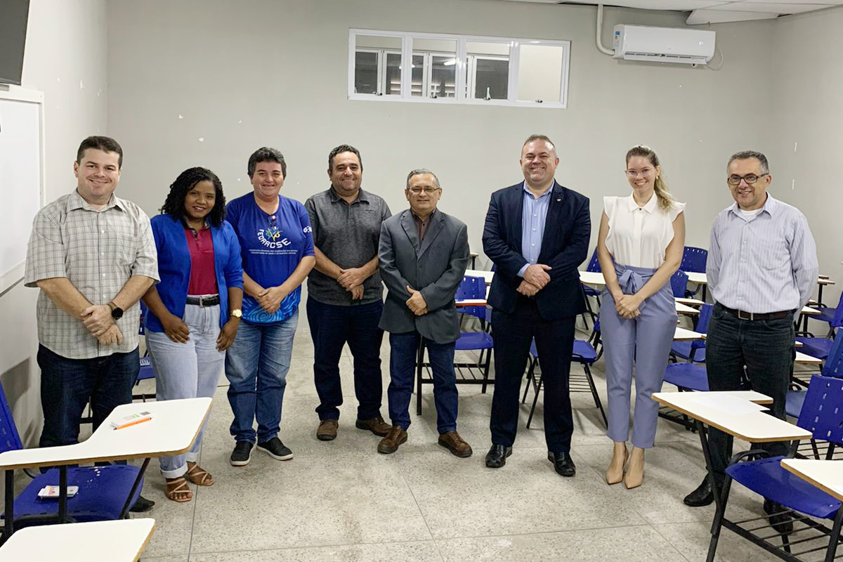 Foto de Duas mulheres e seis homens em pé posam para foto, eles estão numa sala de aula com carteiras azuis e tampos brancos , parede e teto brancos