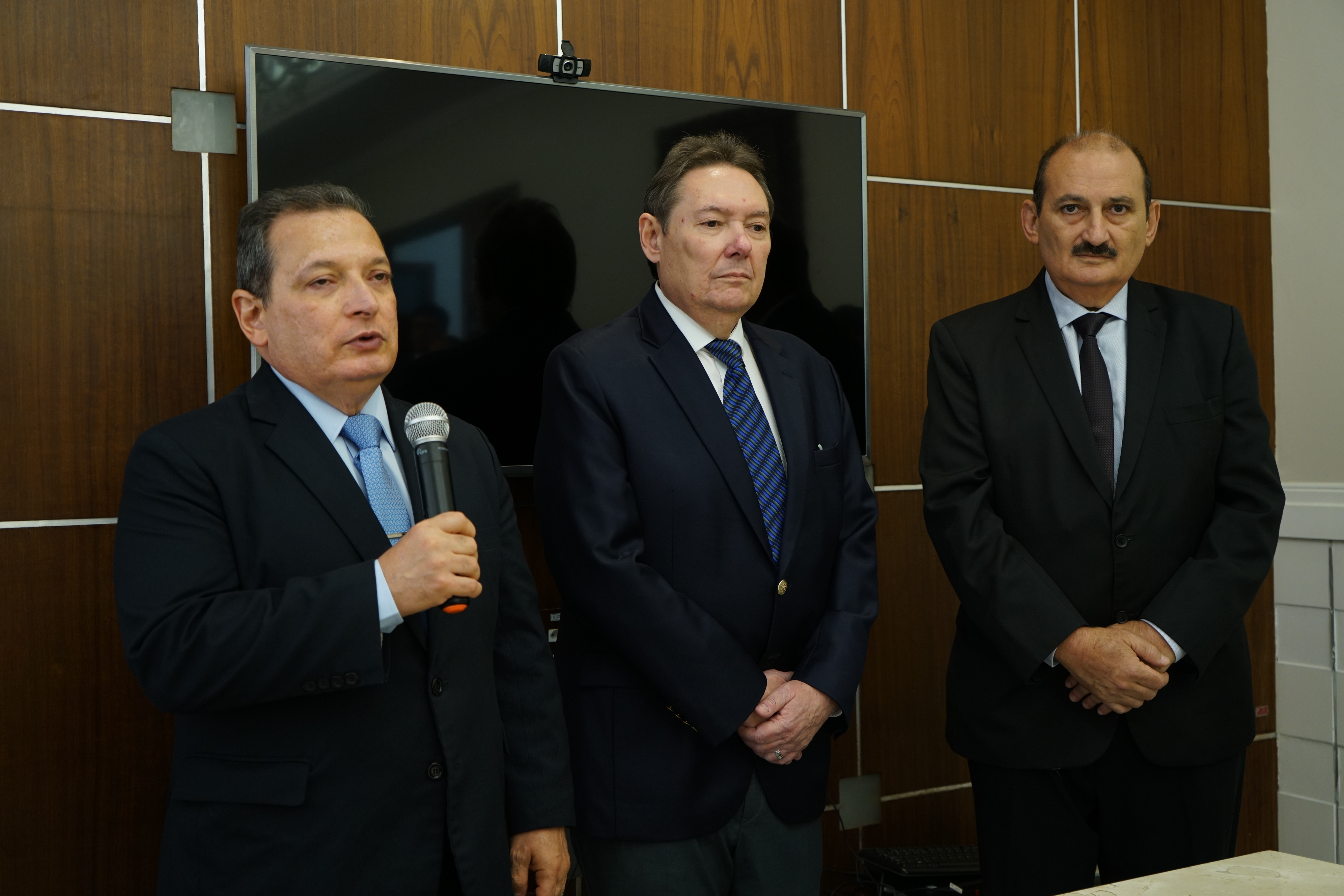 Foto do Presidente do TRT-CE, des. Durval César, empresário Ivens Dias Branco Jr e o desembargador Francisco José Gomes. O presidente está falando ao microfone, enquanto os outros dois estão ao lado escutando e todos de pé. Todos estão de paletó. Atráves deles, tem uma TV e uma parede com revestimento de madeira.