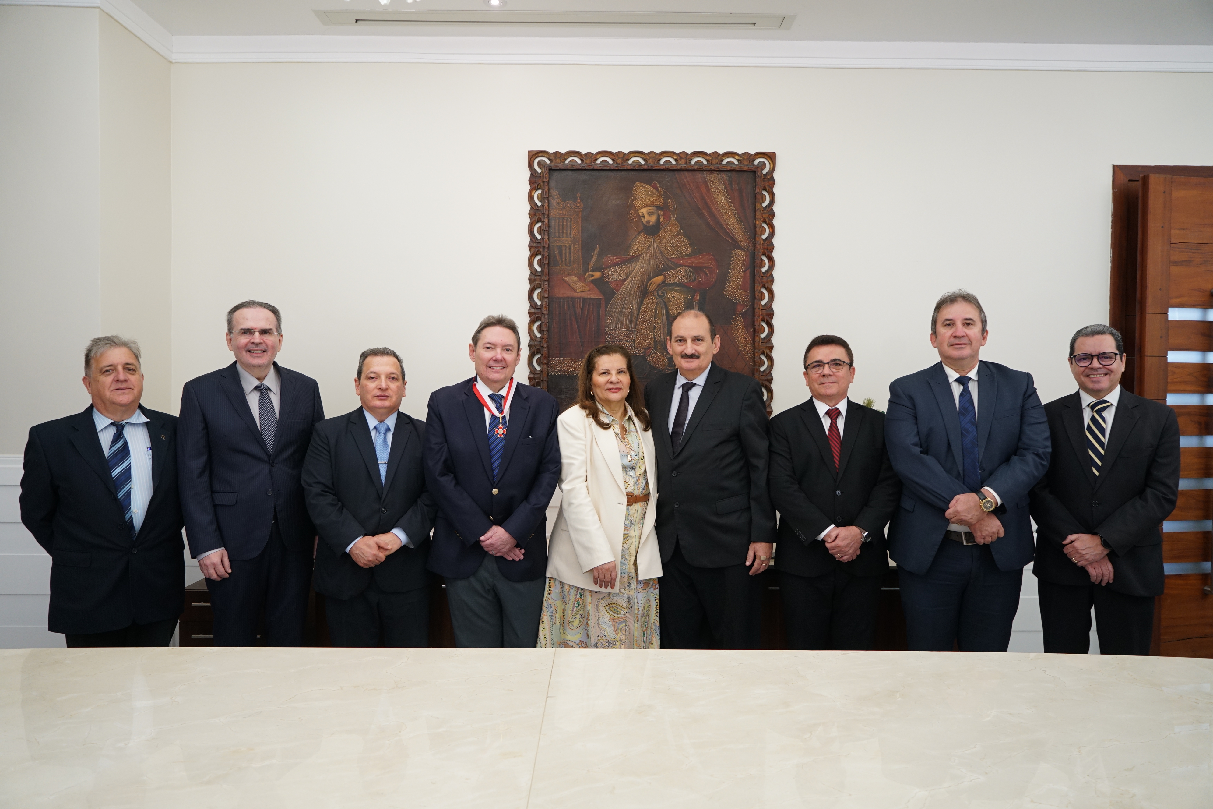 Foto dos Desembargadores do TRT-CE ladeando o empresário Ivens Dias Branco Júnior (com a medalha mais ao centro). Há 8 homens e uma mulher. Eles estão vestino paletós, e a des. Fernanda Uchoa está usando uma roupa clara. Atrás deles, há um quadro com uma pintura de  um homem escrevendo. Parece ser um representante da Igreja Católica pelas vestes e chapéu