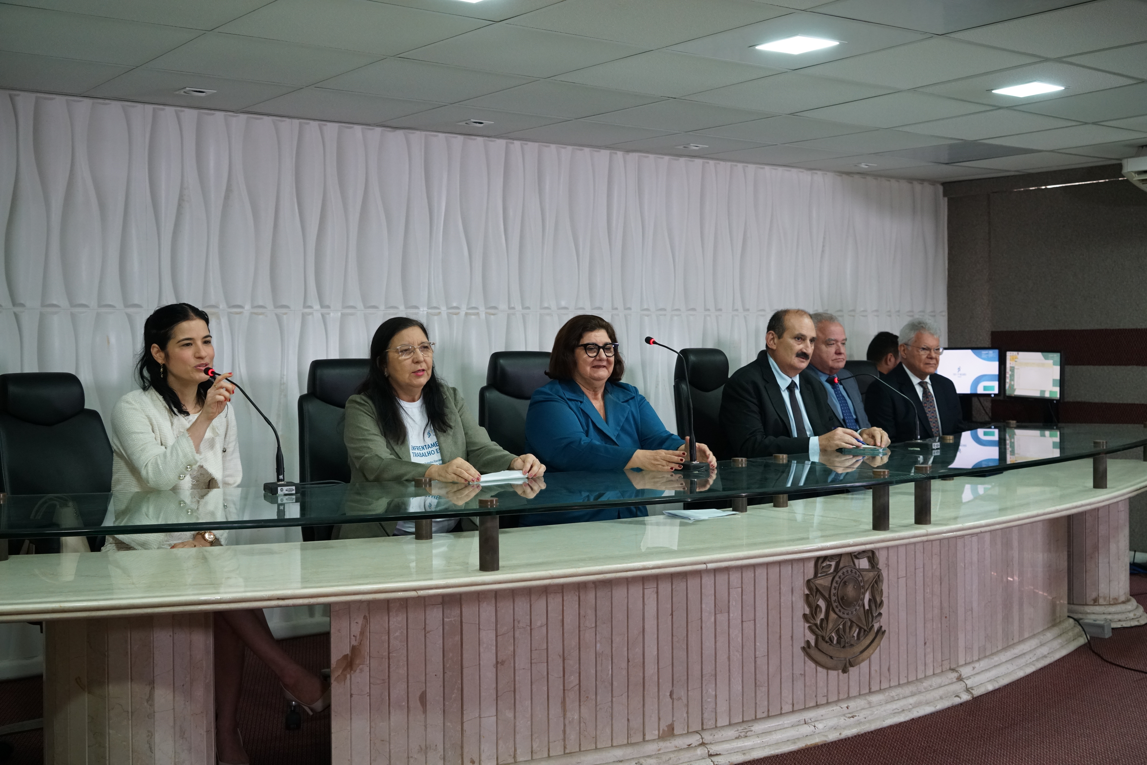 Foto da mesa do Seminário onde estão sentados a gestora regional do Programa Trabalho Seguro, desa. Regina Gláucia Cavalcante; o gestor regional do Programa de Enfrentamento ao Trabalho Escravo, des. Francisco José Gomes da Silva; a coordenadora do evento Francinete Giffoni; o presidente da Amatra7, juiz Hermano Queiroz Júnior; a procuradora-chefe do Ministério Público do Trabalho, Geórgia Silveira Aragão; e o conselheiro do Conselho Regional de Medicina (CRM), Anastácio de Queiroz Sousa. Nesse momento, a procuradora do trabalho Geórgia Aragão está falando no microfone, enquanto o restante dos integrantes da mesa está olhando para o auditório. Todos estão vestidos formalmente.