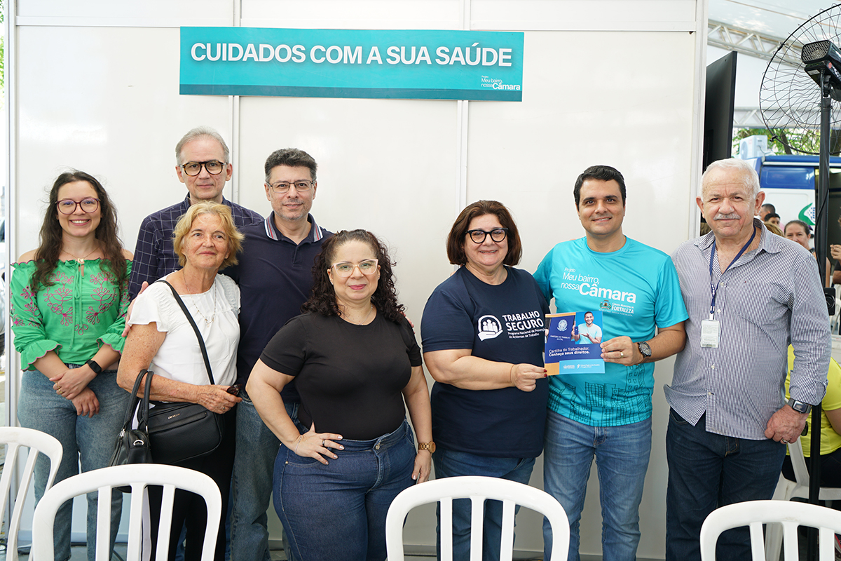 Oito pessoas em pé num estande branco, a mulher no centro segura uma cartilha com o título Cartilha do Trabalhador