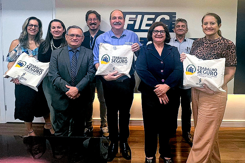 quatro homens e quatro mulheres em pé posam para foto, em frente a uma parede branca com o letreiro FIEC. Três deles seguram uma bolsa de lona com o logotipo Programa Trabalho Seguro