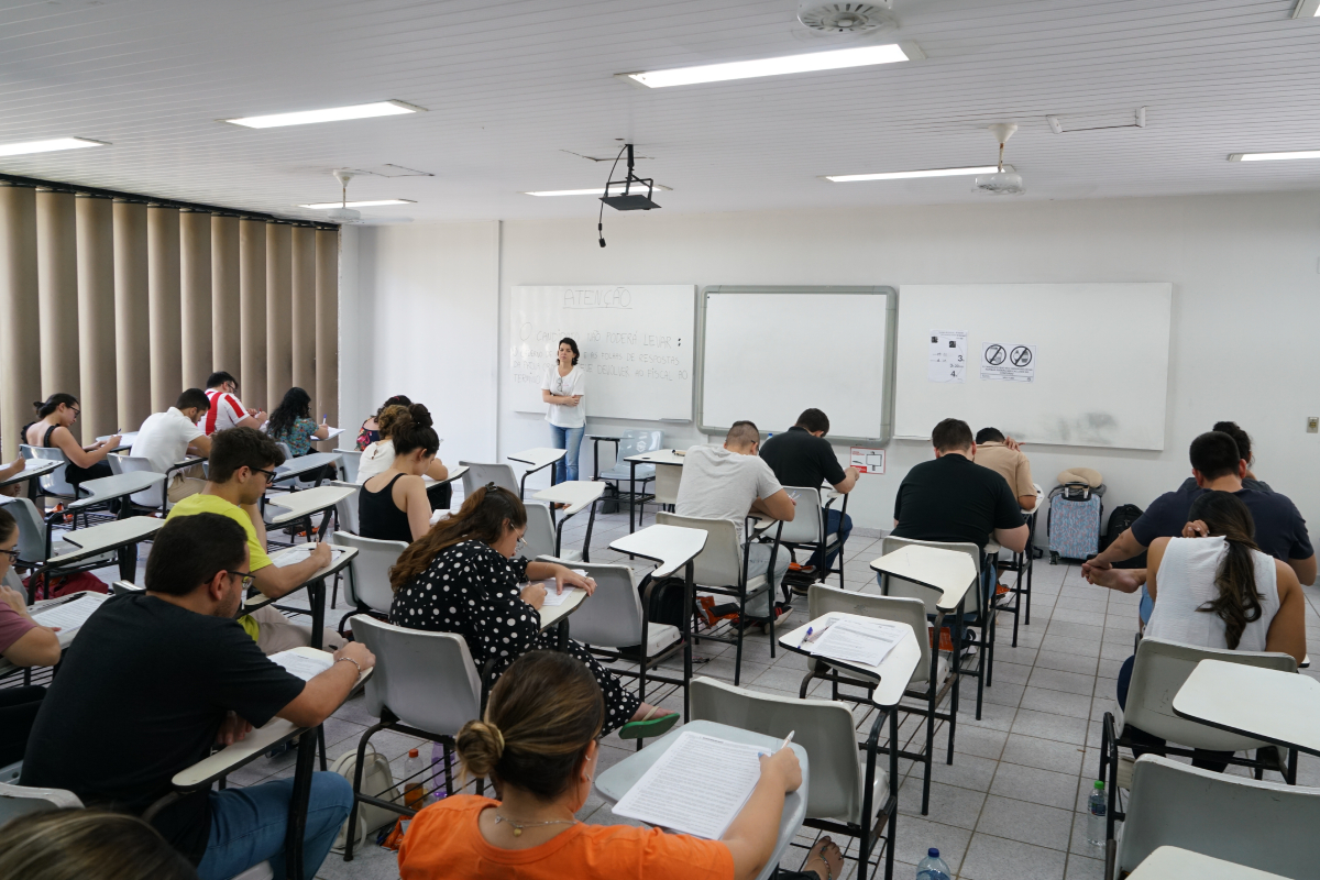 Foto de pessoas sentadas em uma sala de aula. Elas escrevem e lêem as provas que estão em cima das carteiras. Em pé, à frente delas, está uma pessoa em pé com a identificação no peito Fiscal