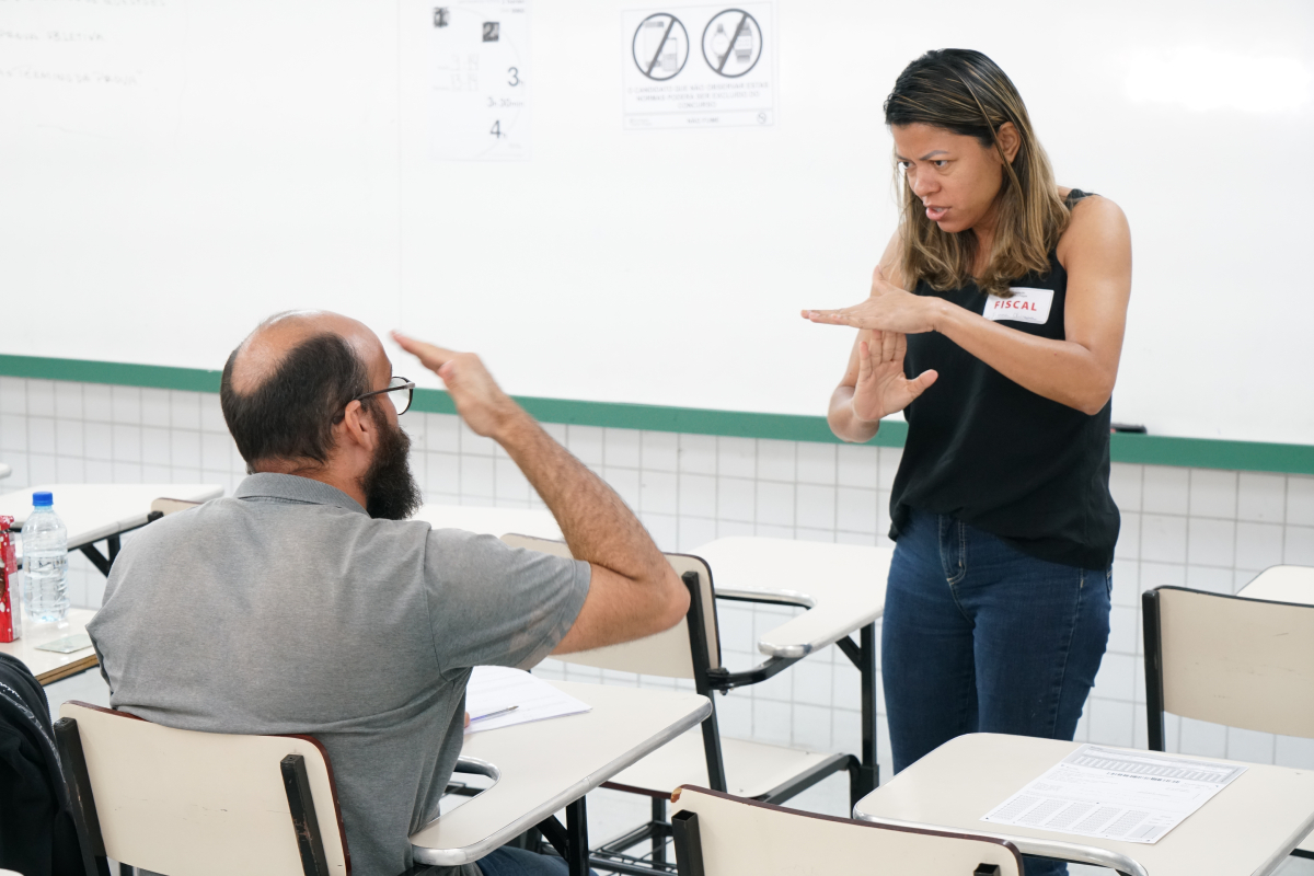 Homem sentado na carteira da sala de aula com a prova na frente. Ele olha para uma mulher identificada como Fiscal. Ela está gesticulando um sinal de Libras para ele.