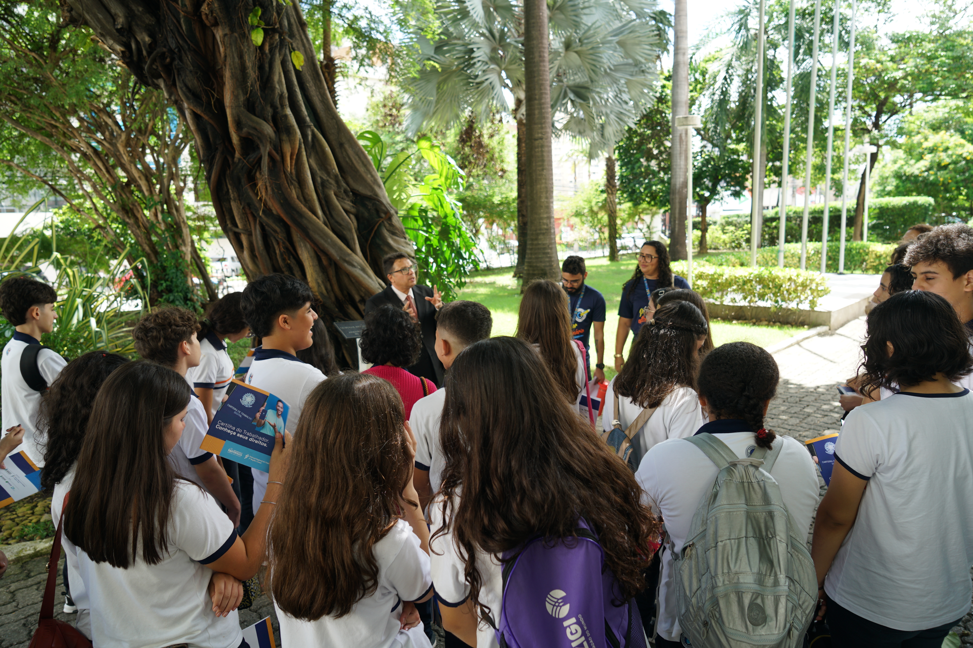 Foto tirada no jardim da casa sede do TRT. Há vários estudantes fardados ouvindo a explicação do cerimonialista do TRT, Antônio Carlos Santiago. Eles estão sob a sombra de uma grande árvore. 